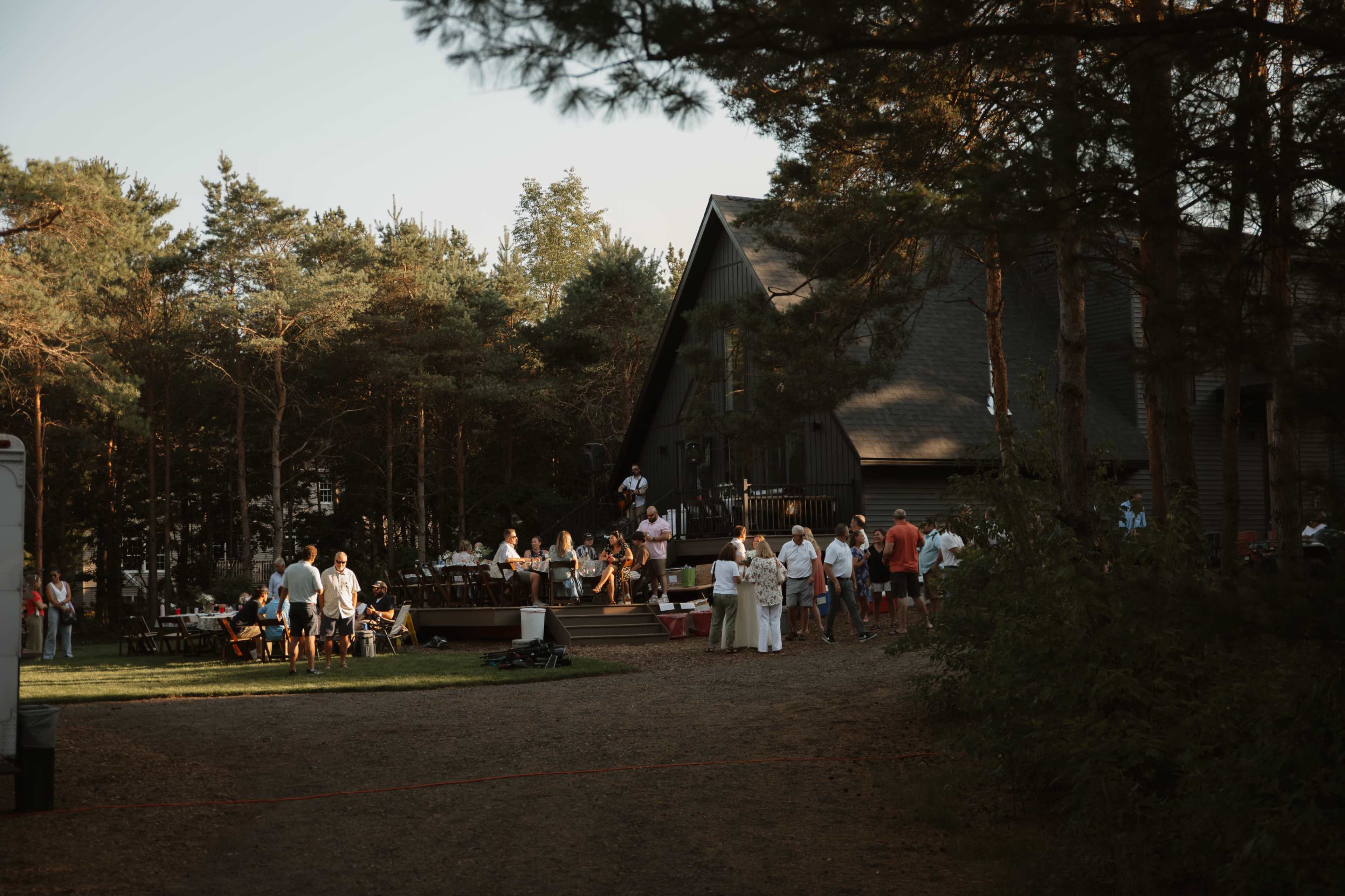 A gathering of people is taking place outside a large, dark-colored building surrounded by trees.