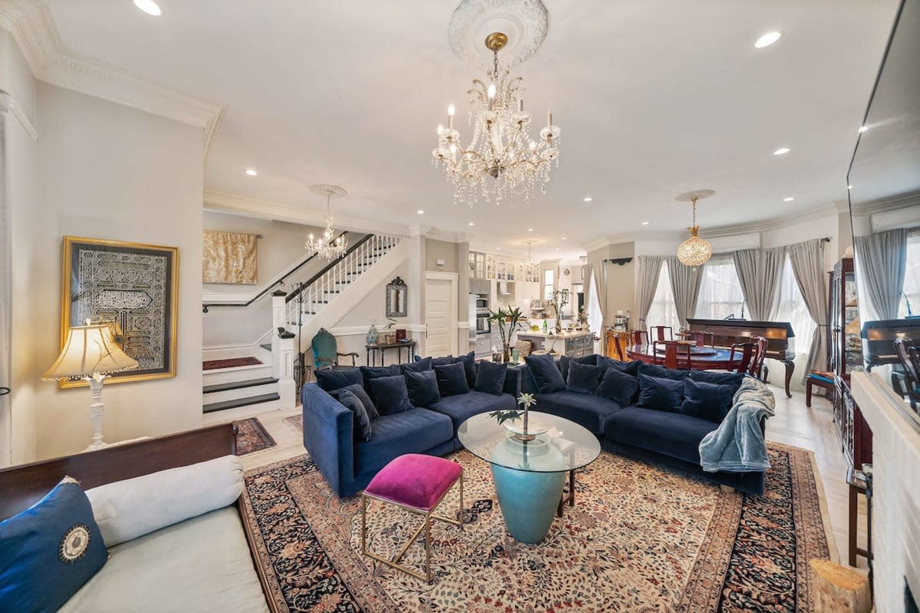 A well-lit living room with a large navy blue sectional couch, a glass coffee table, a chandelier, and a staircase leading to the upper level.