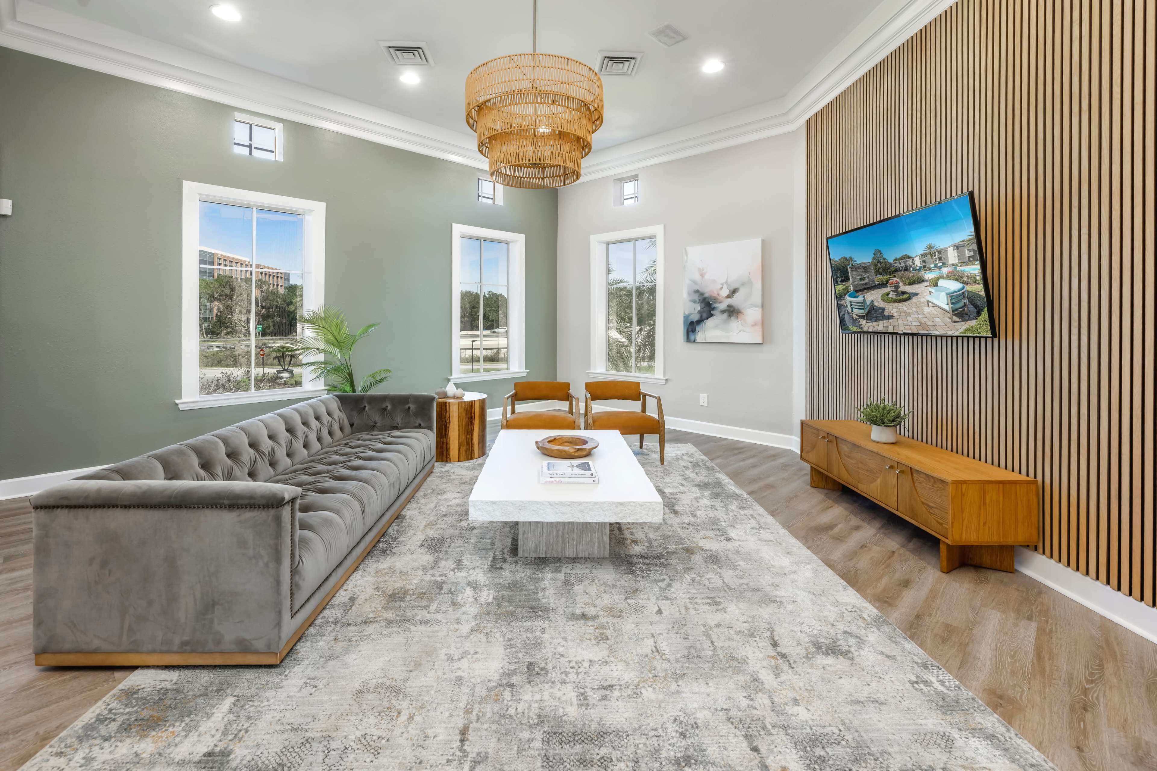 A modern living room features a gray velvet sofa, a marble coffee table, a wooden TV stand, and large windows allowing natural light.