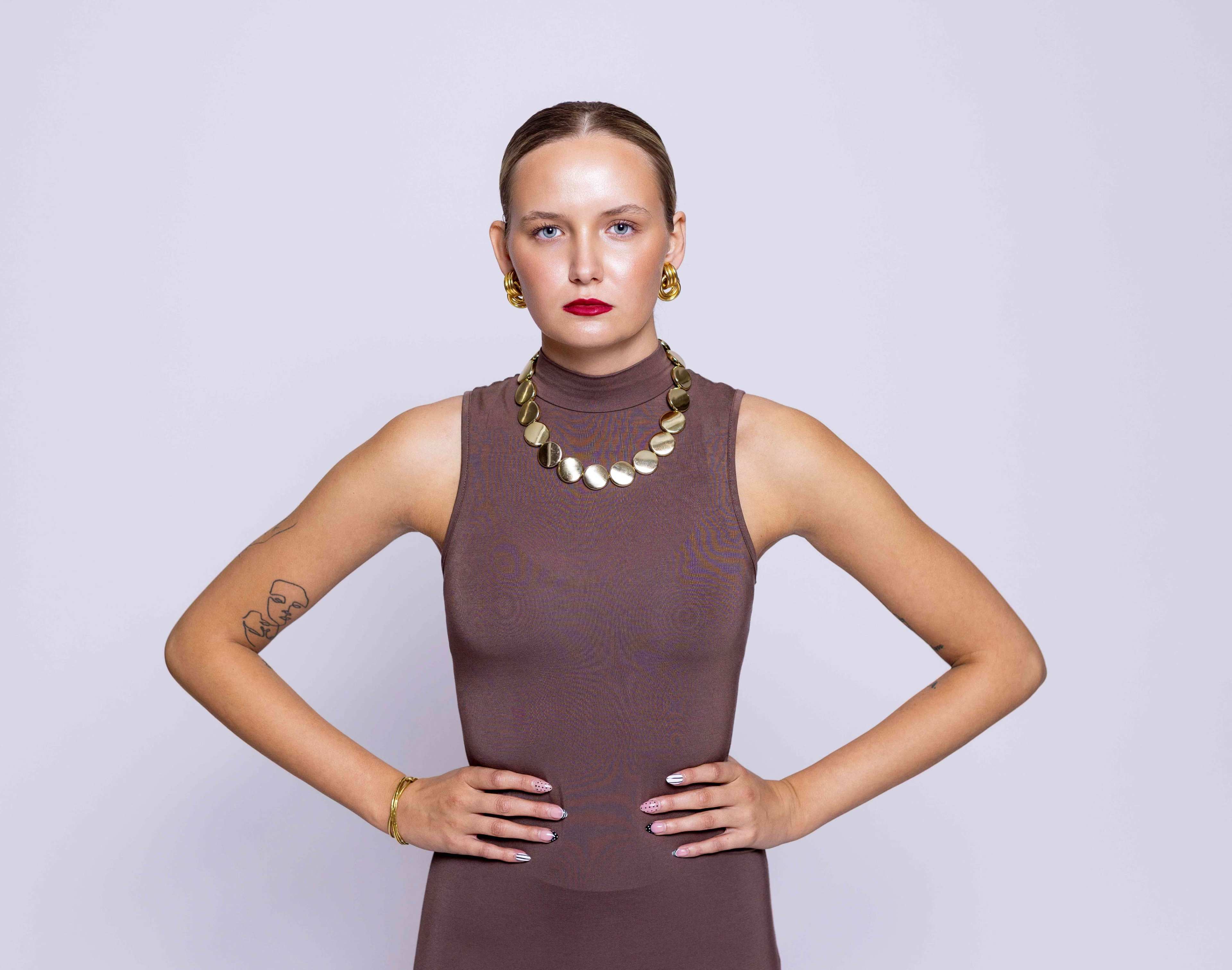 A model poses with a serious expression, wearing a sleeveless brown top and statement jewelry against a plain background.