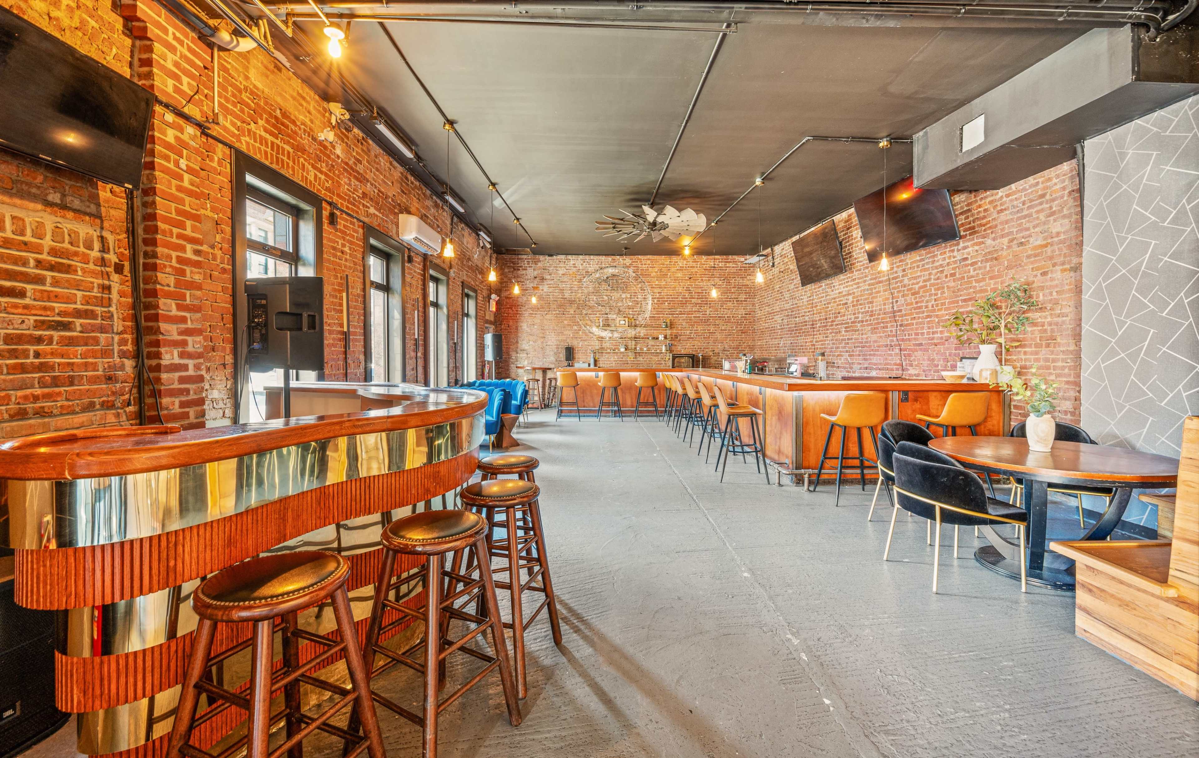 The interior of a bar features exposed brick walls, wooden furniture, and pendant lighting, with a curved wooden bar in the foreground.