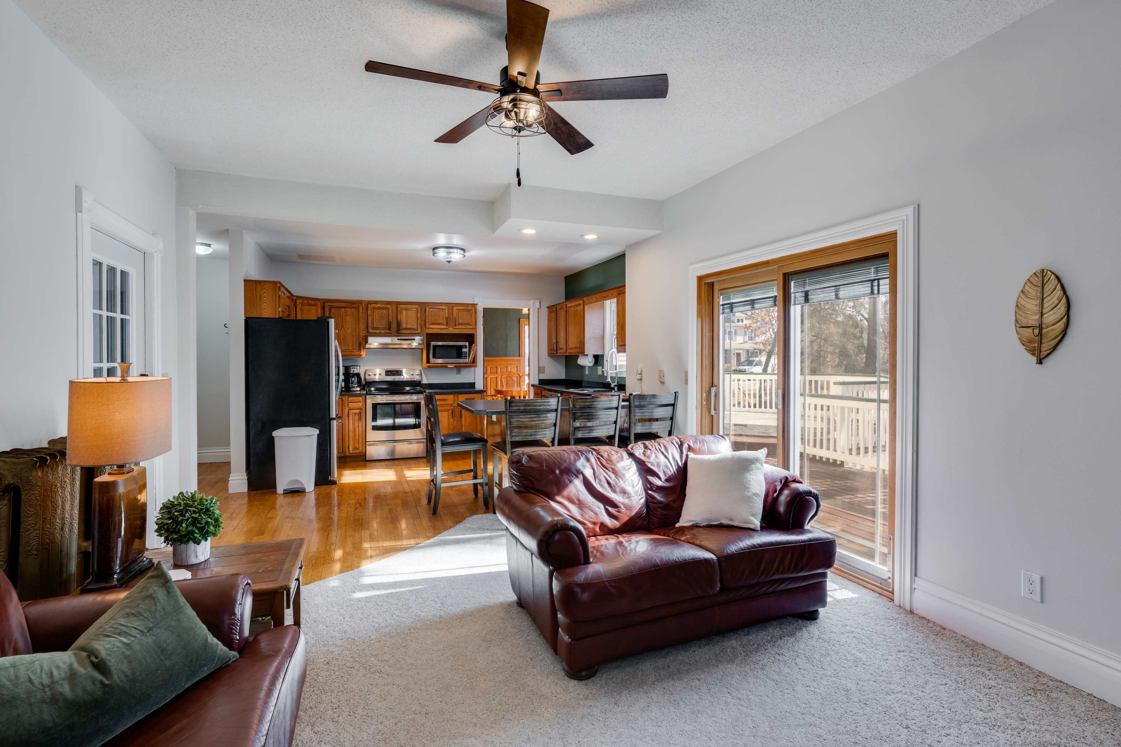 The image shows a living room with a leather couch, a ceiling fan, and a view of a kitchen and dining area in an open floor plan.