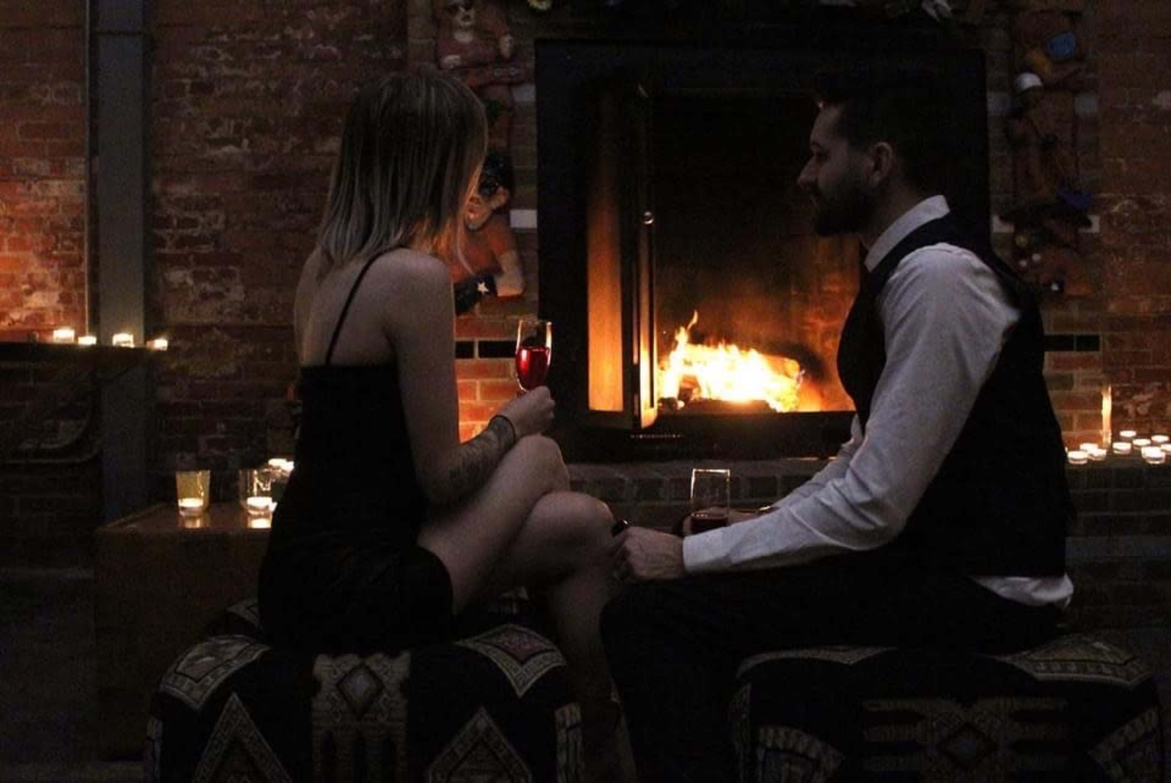 A man and woman sit on a low table in front of a fireplace, each holding a drink.