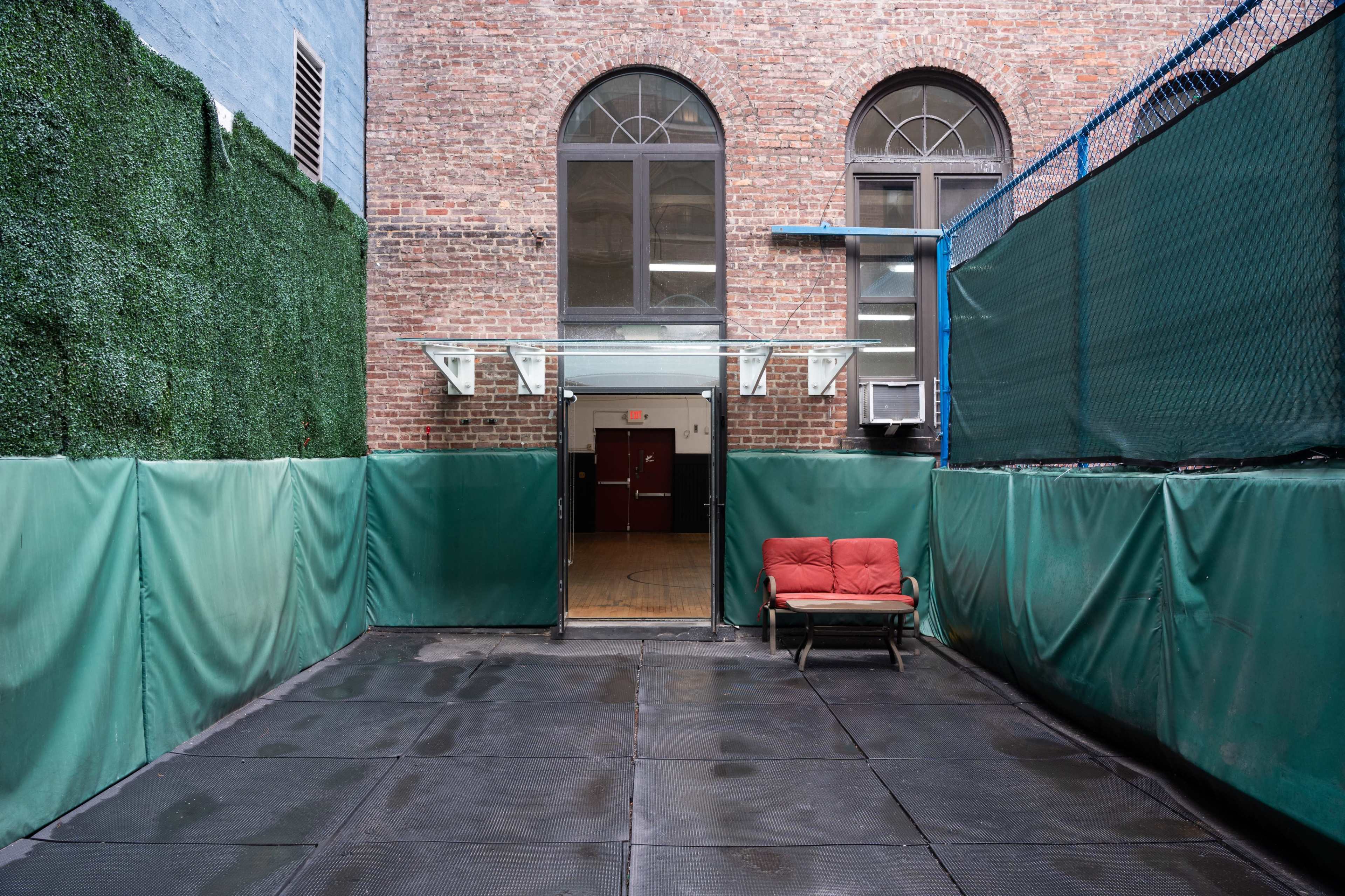 The image shows a small, enclosed outdoor area with a red couch and a door leading to a brick building.