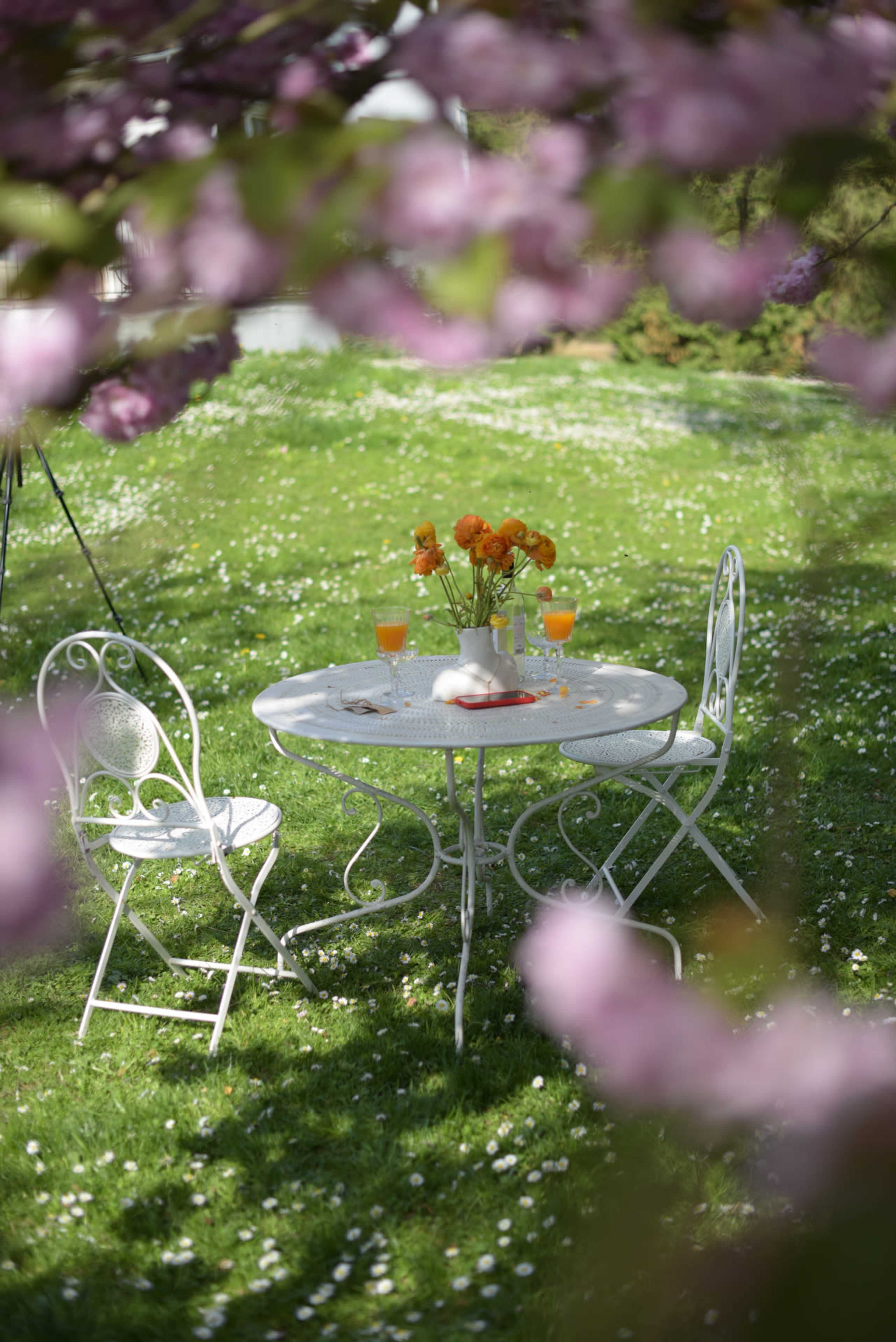 A white metal table with two chairs is set on a grassy area adorned with fallen petals, featuring a centerpiece of orange flowers and drinks.