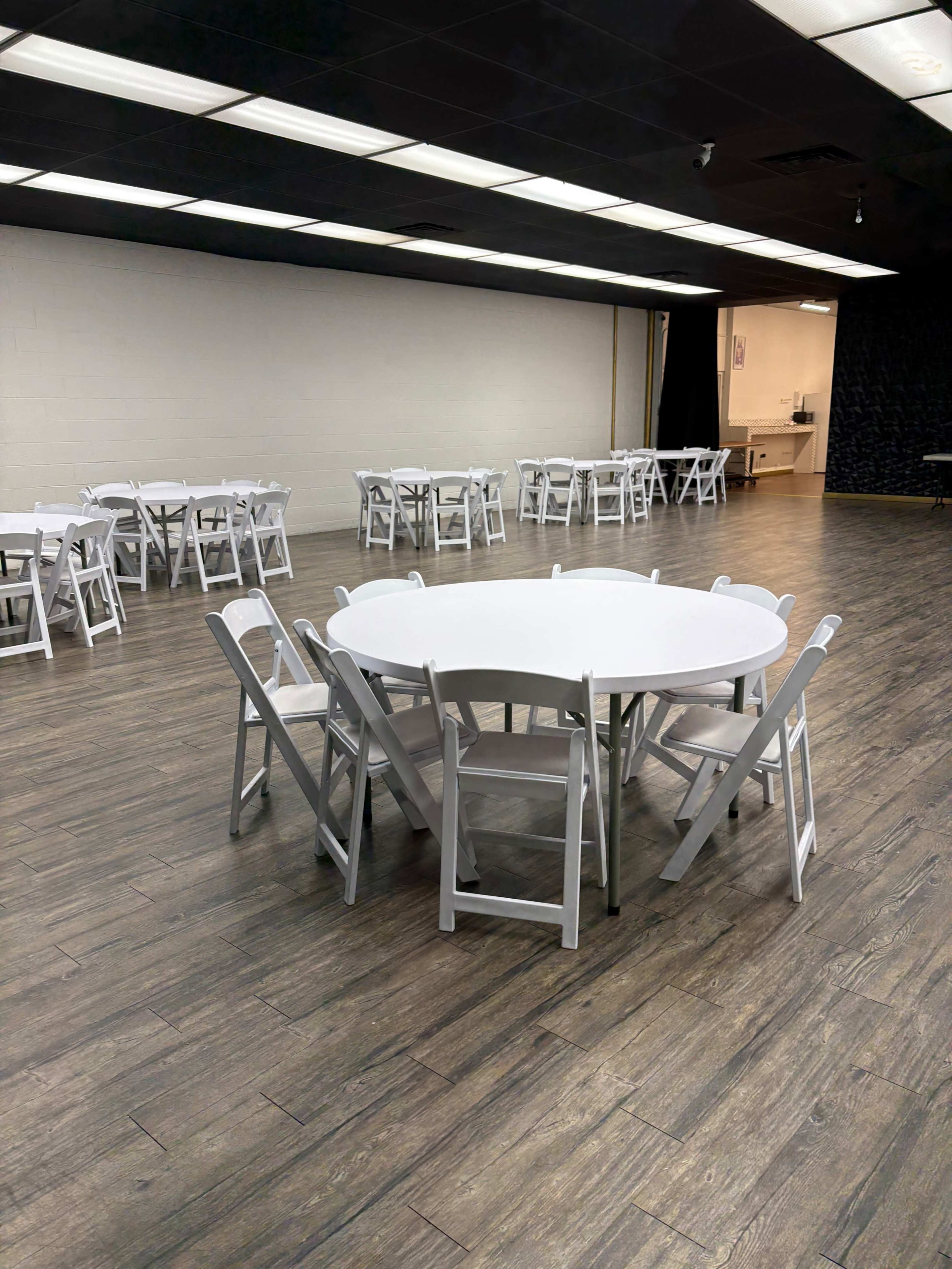 The image shows a spacious room with several tables set up in a circle, surrounded by white folding chairs on a wooden floor.
