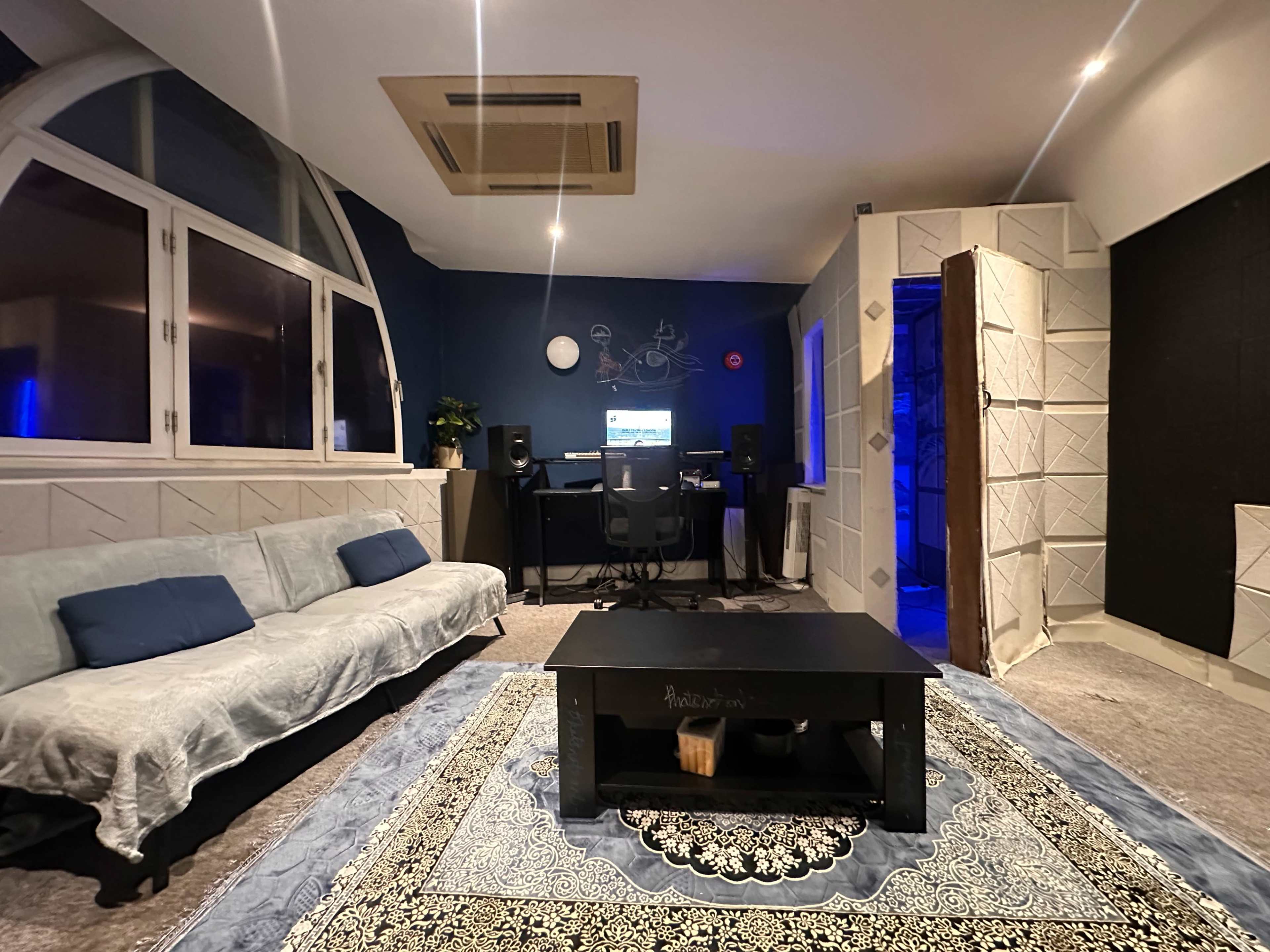 The image shows a cozy room featuring a light blue sofa, a coffee table, and a desk with computer equipment in the corner, all under soft lighting.