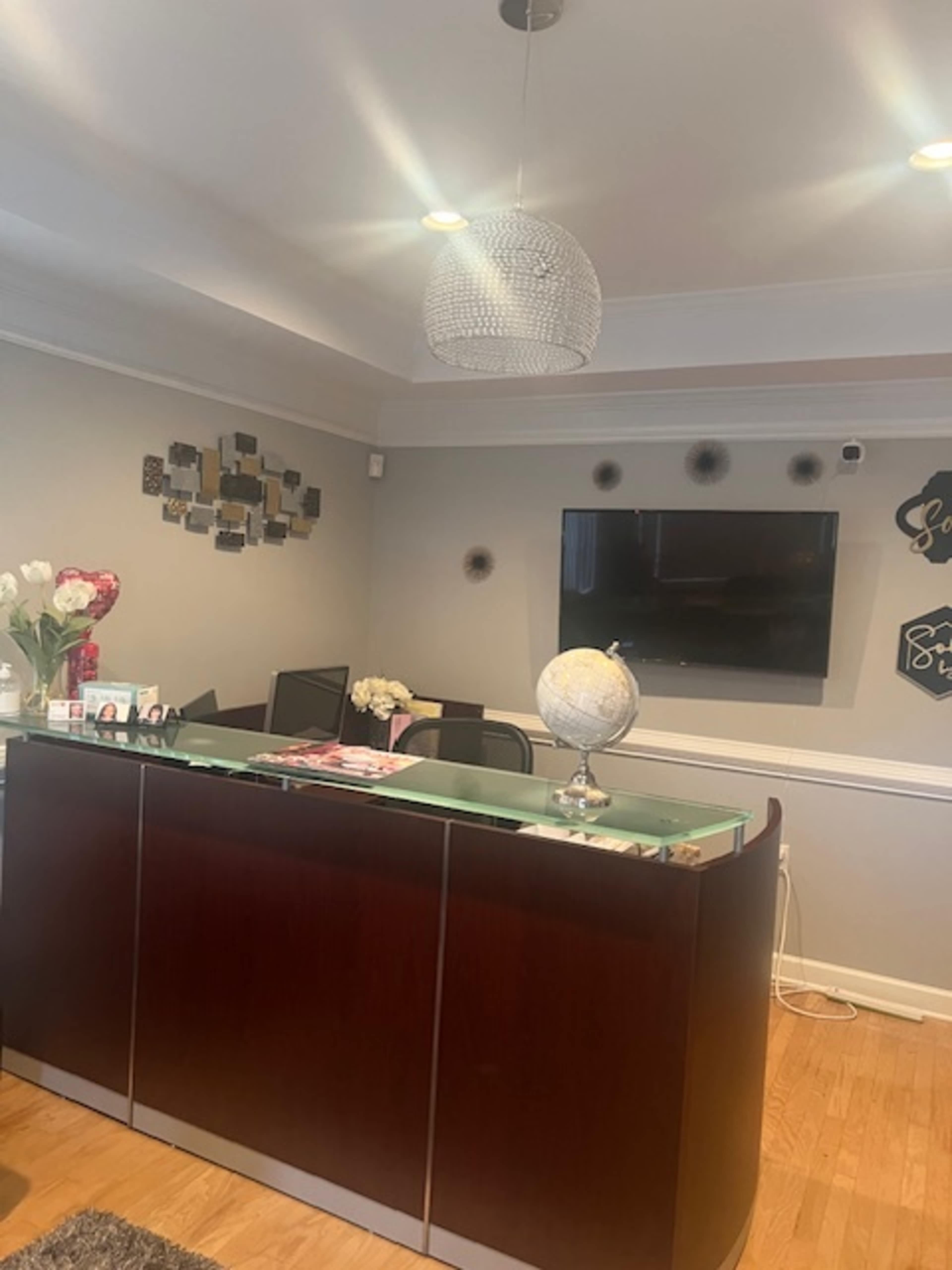 A reception area featuring a wooden front desk with a glass top, a flat-screen TV mounted on the wall, and decorative wall art along with flowers on the counter.