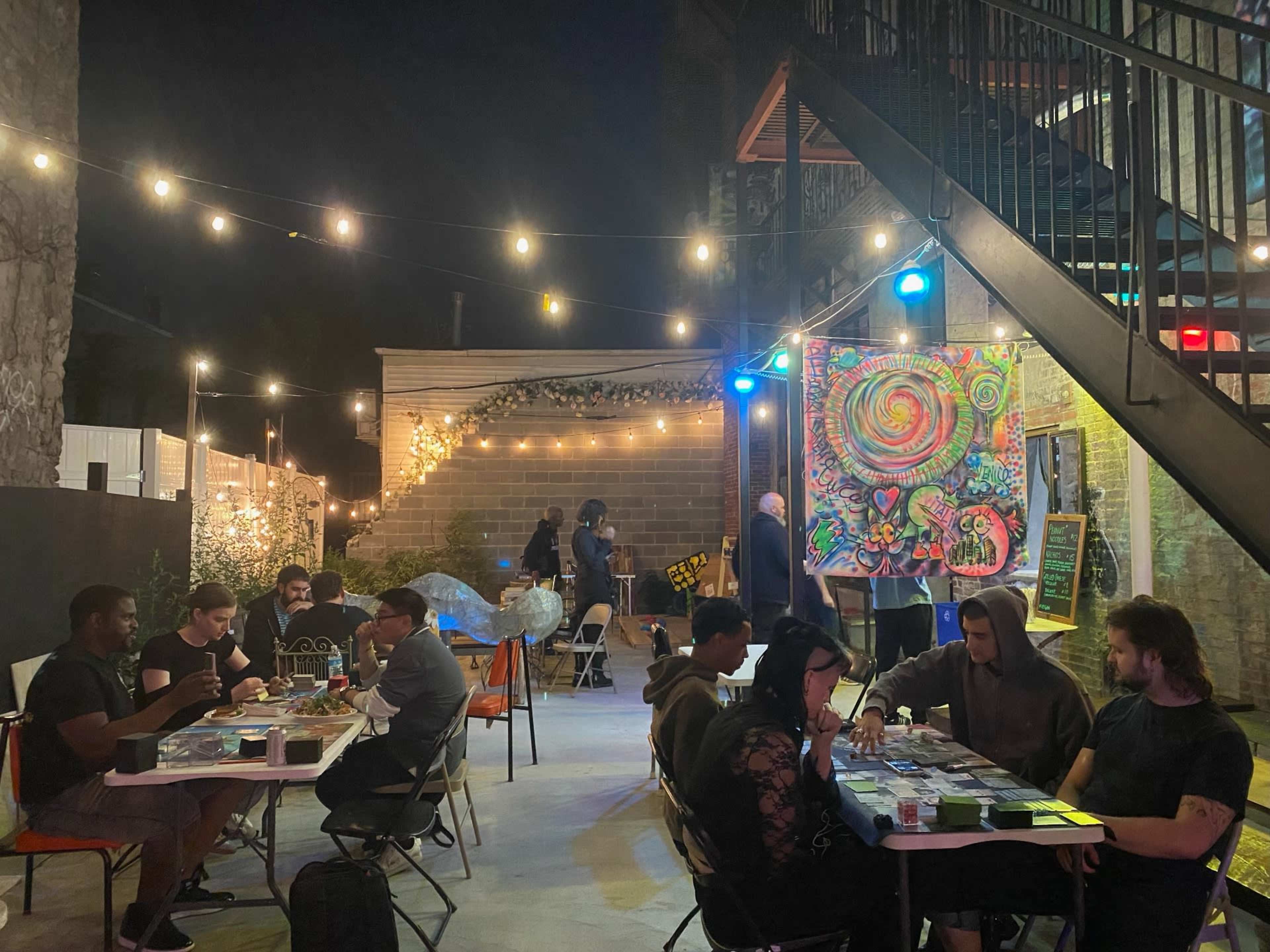 A group of people is seated around tables in an outdoor area at night, engaged in various activities while string lights illuminate a colorful mural and the surrounding space.