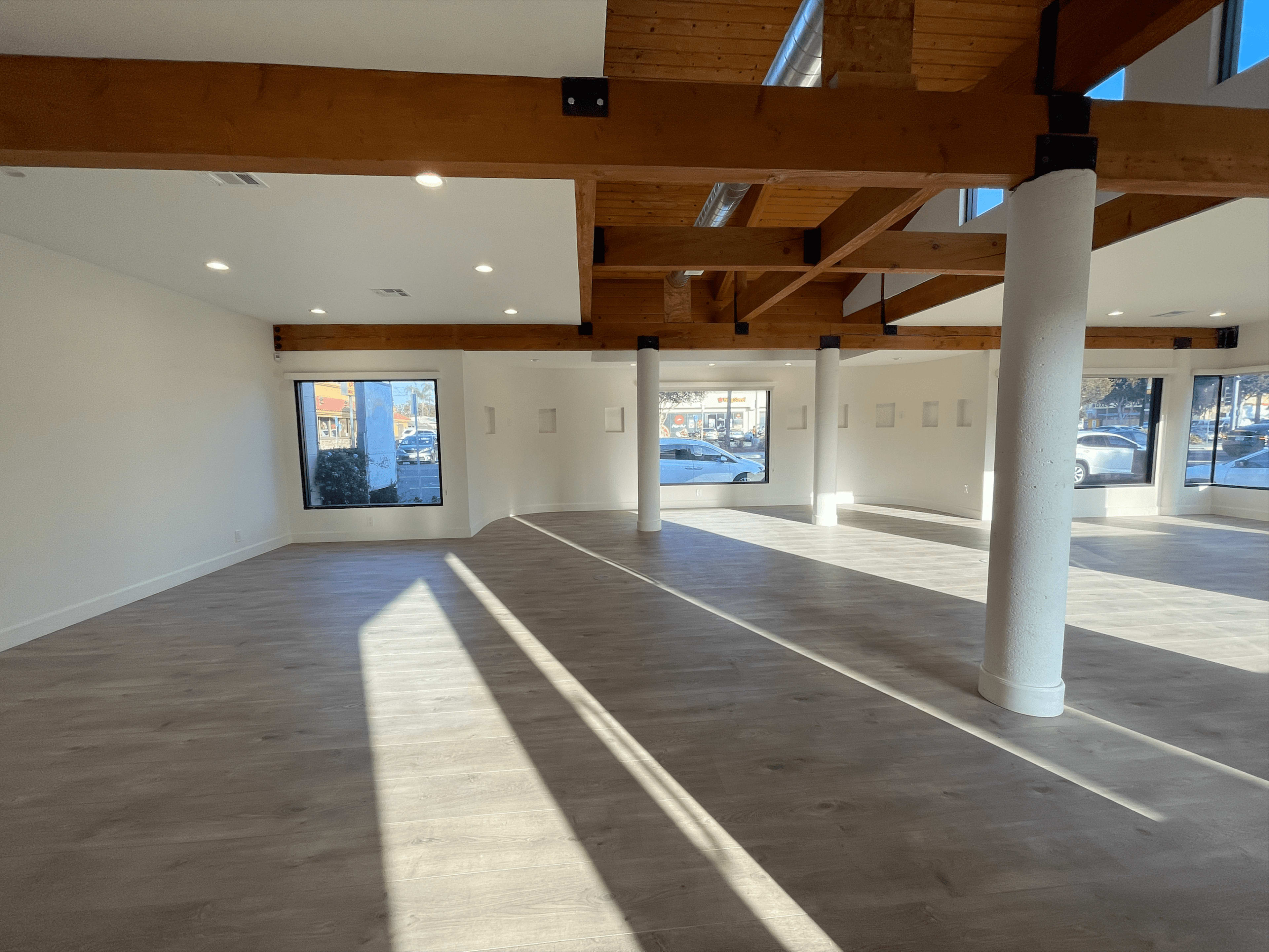 The image shows an empty interior space with wooden beams, large windows, and light-colored flooring.