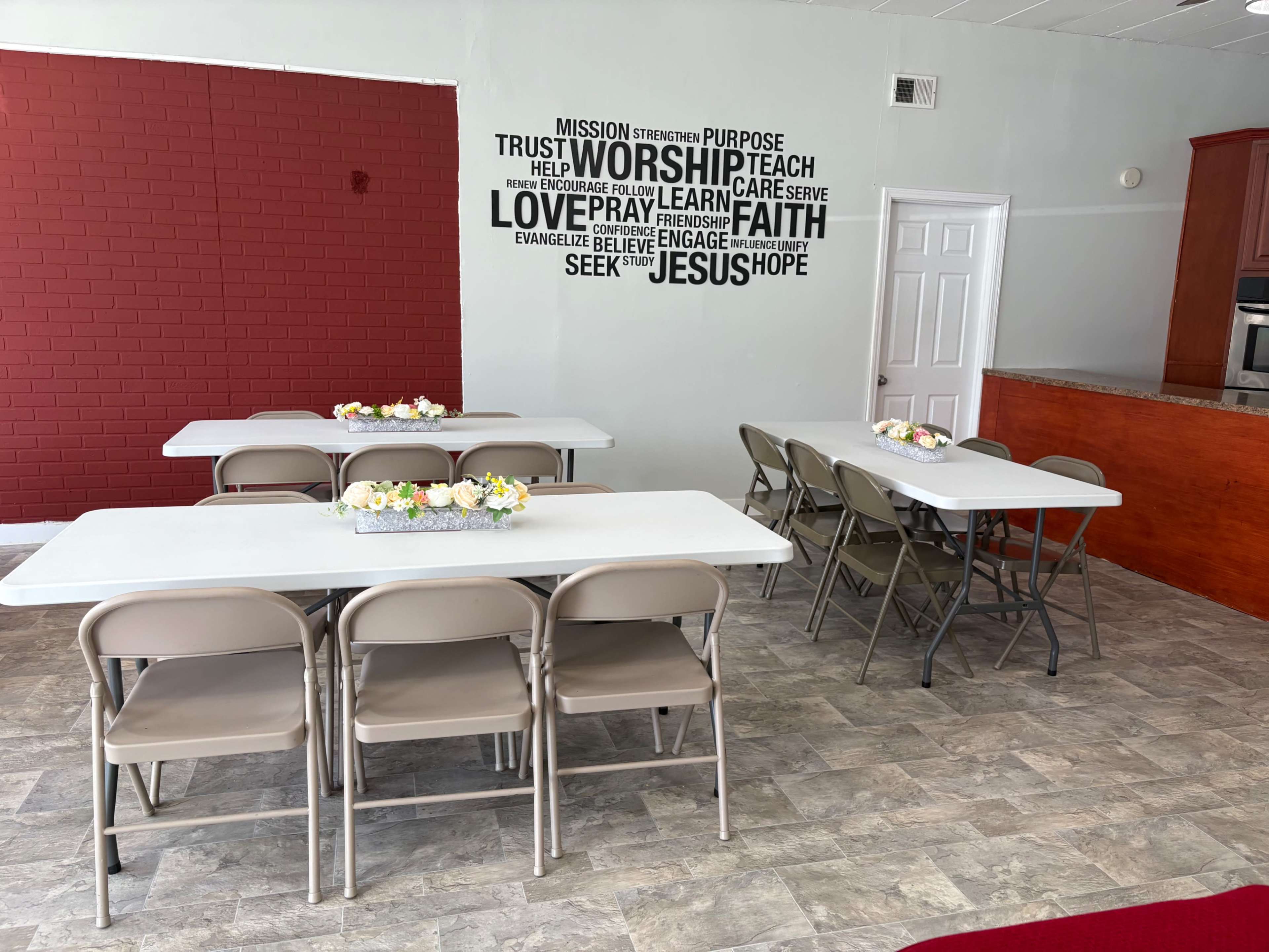 The image shows a community room with several tables set up, flowers as centerpieces, and a motivational wall mural featuring words like "Worship," "Faith," and "Hope."