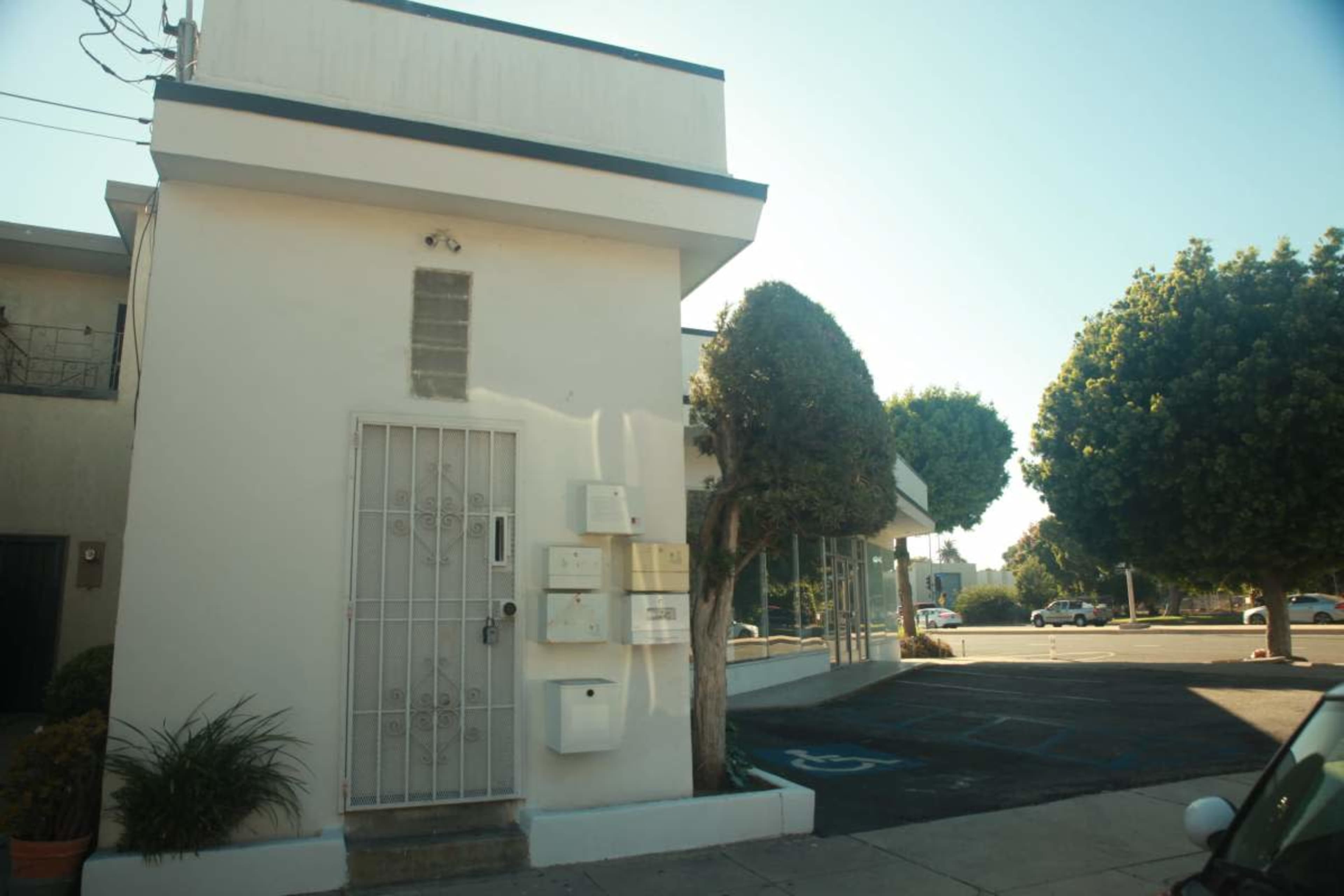 The image shows a white two-story building with a gated entrance, featuring a mailbox and trees in front, situated in a parking lot.