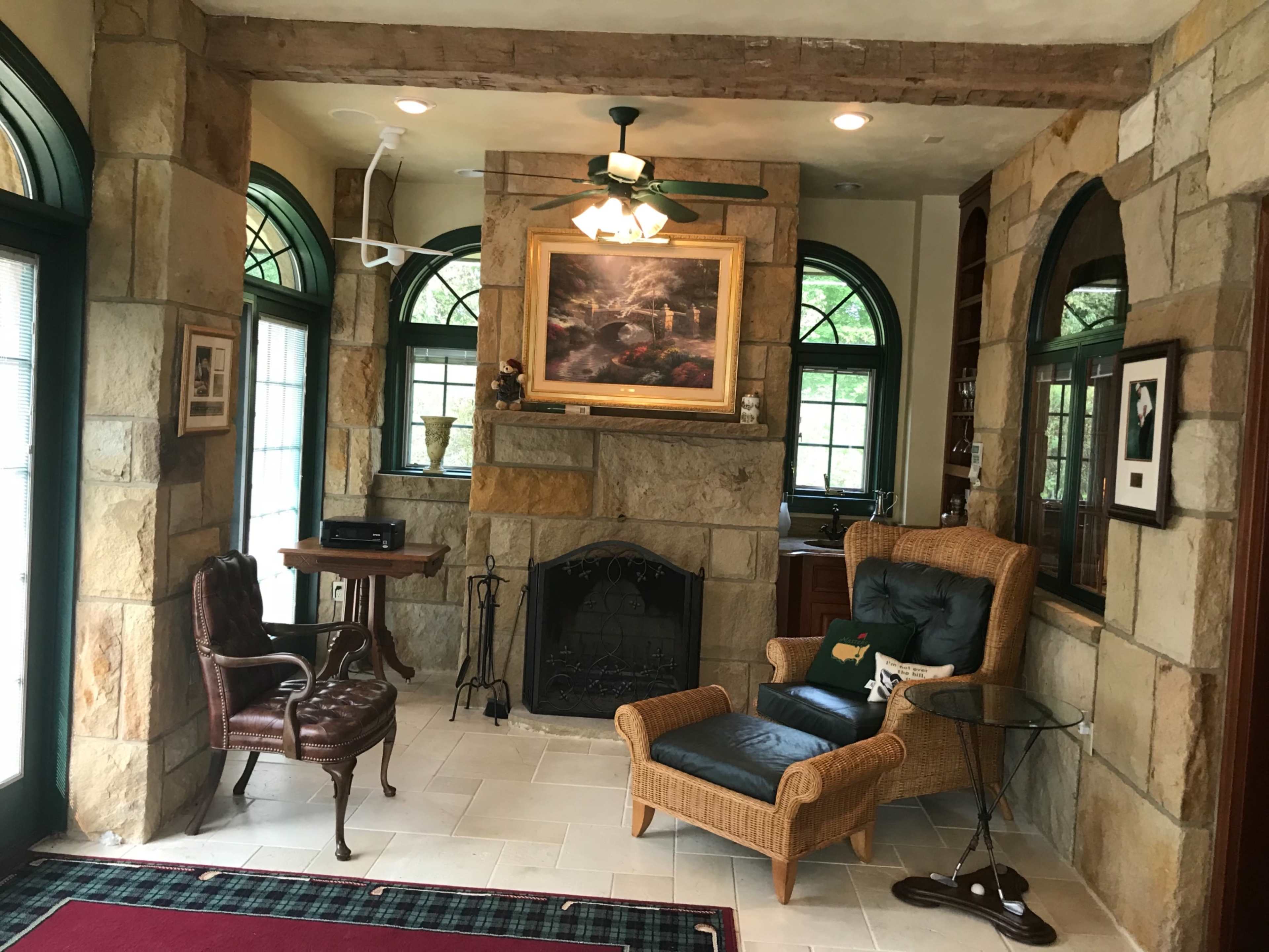 The room features a stone wall, a fireplace, a wicker armchair, a wooden chair, and large windows.