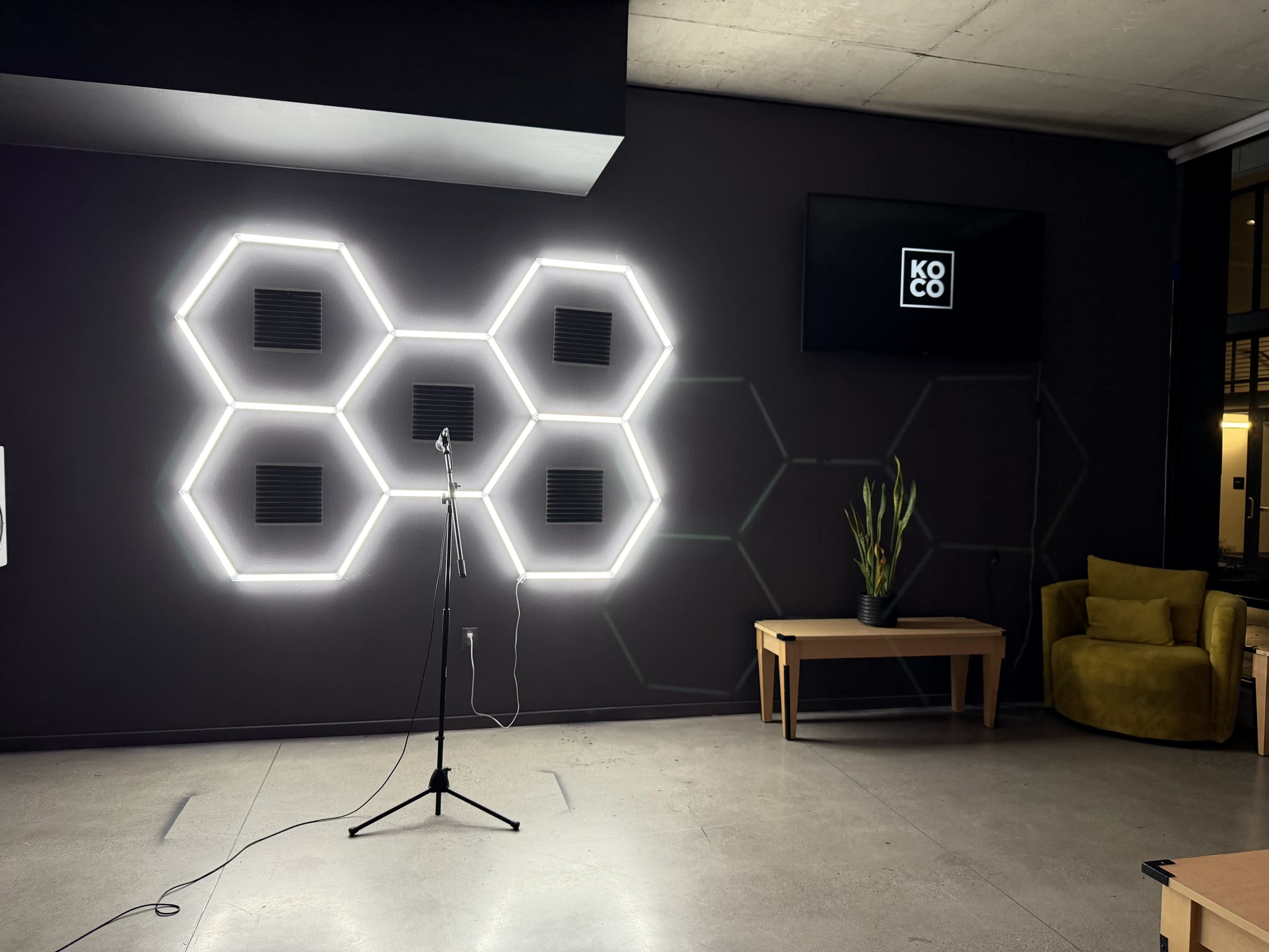 The image shows a modern interior space featuring a hexagonal neon light design on the wall, a microphone stand, a plant, a coffee table, and a green chair.