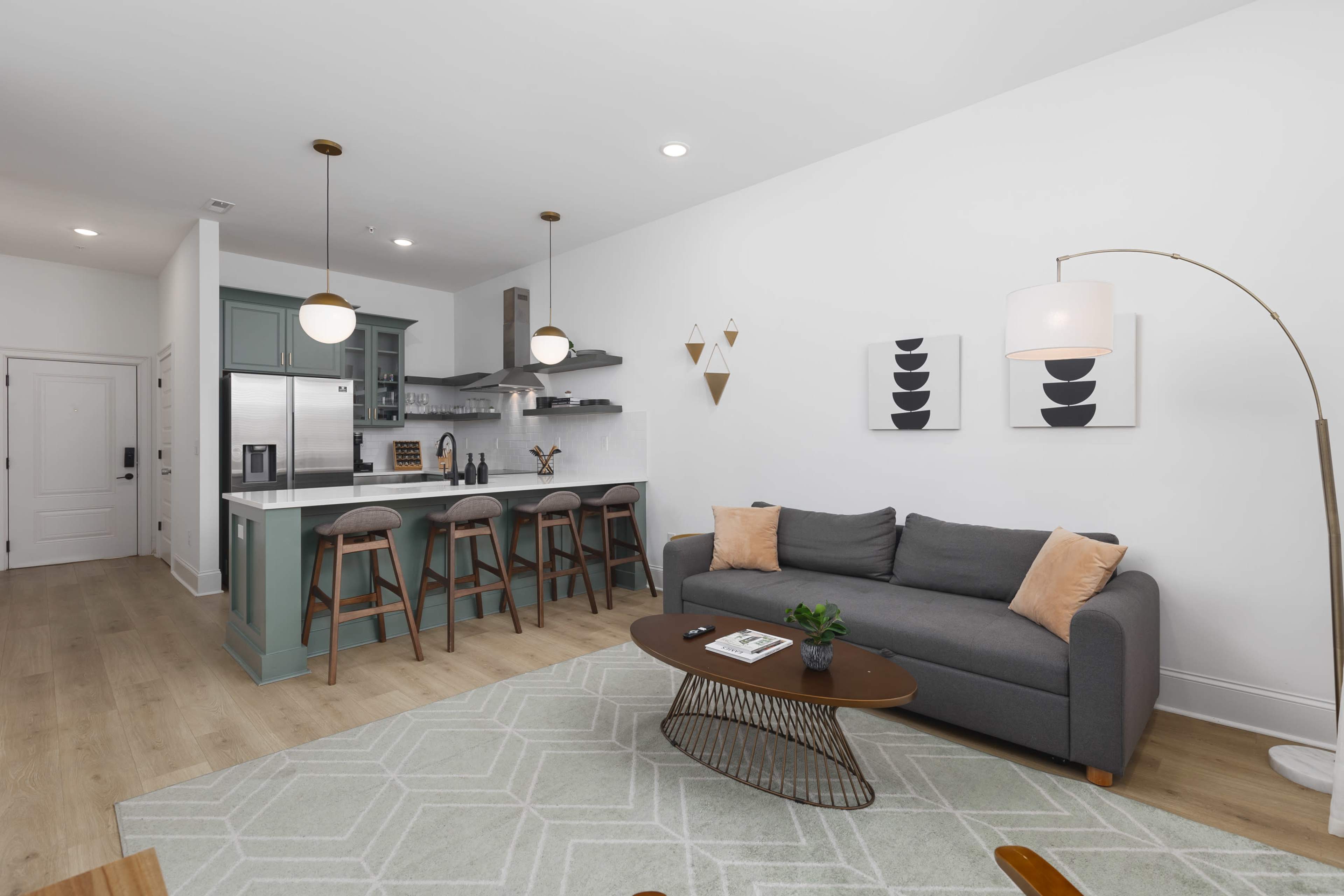 The image shows a modern living room with a gray couch, a wooden coffee table, and a kitchen area featuring bar stools and green cabinetry.