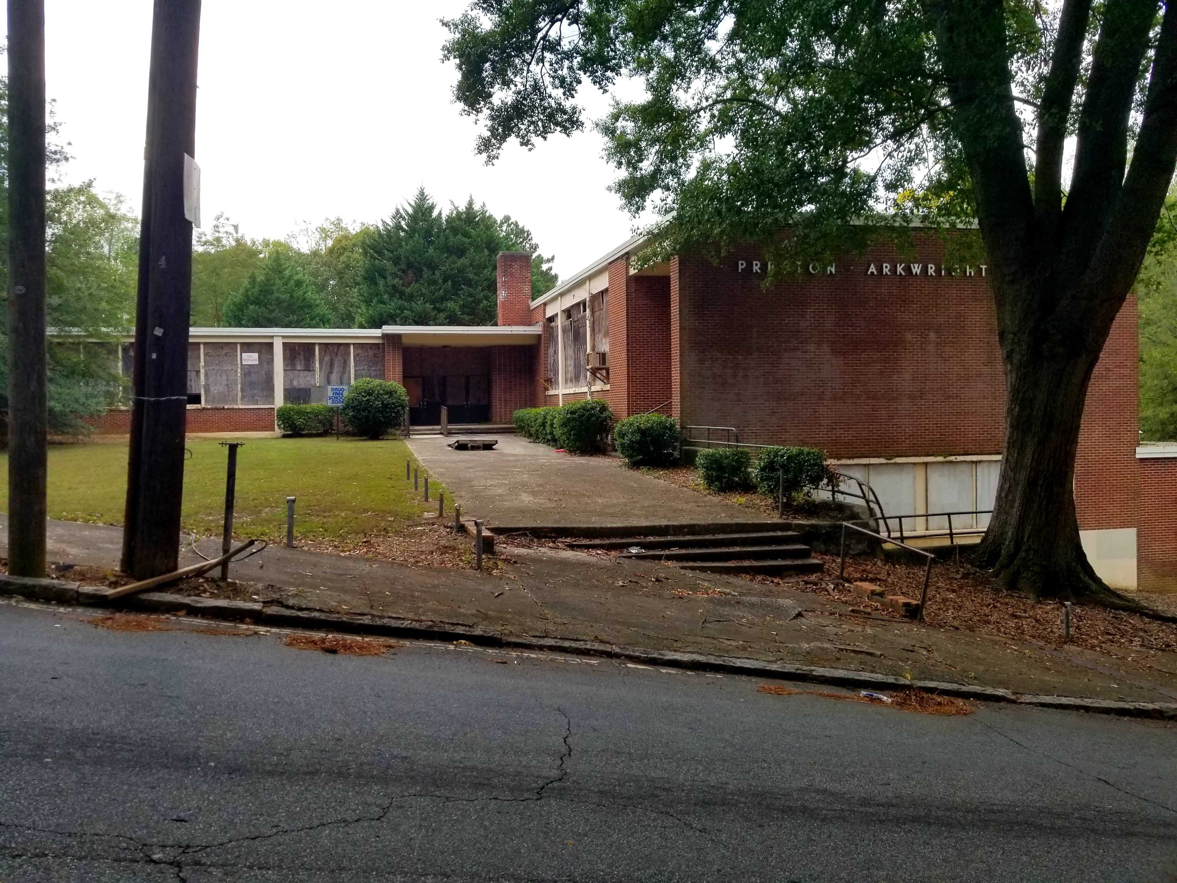 A brick building with large windows, set on a sloped lawn and bordered by trees, with a staircase leading up to the entrance.