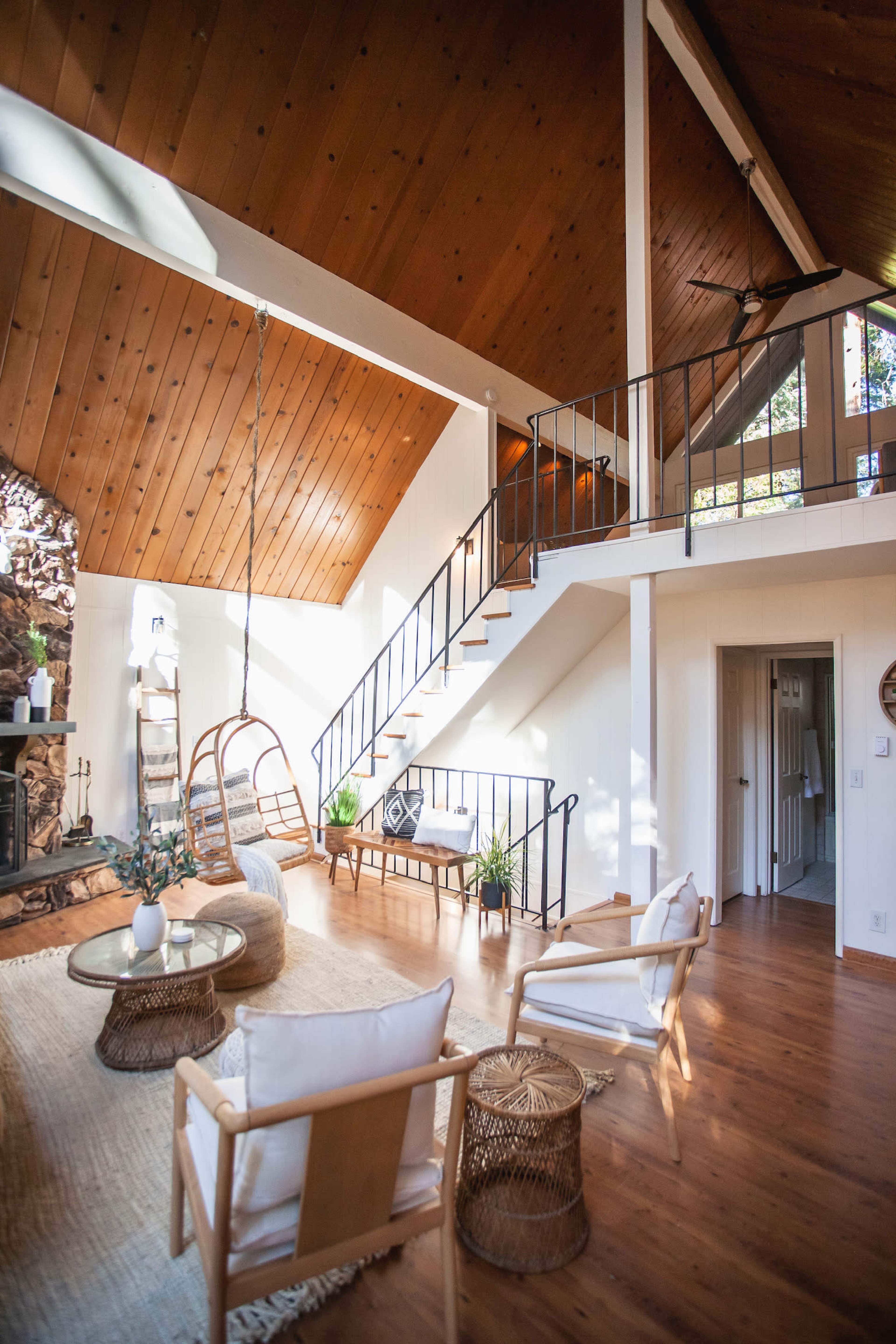 The image shows a bright living room with high wooden ceilings, a staircase, and a cozy seating area featuring a hanging chair and natural decor.