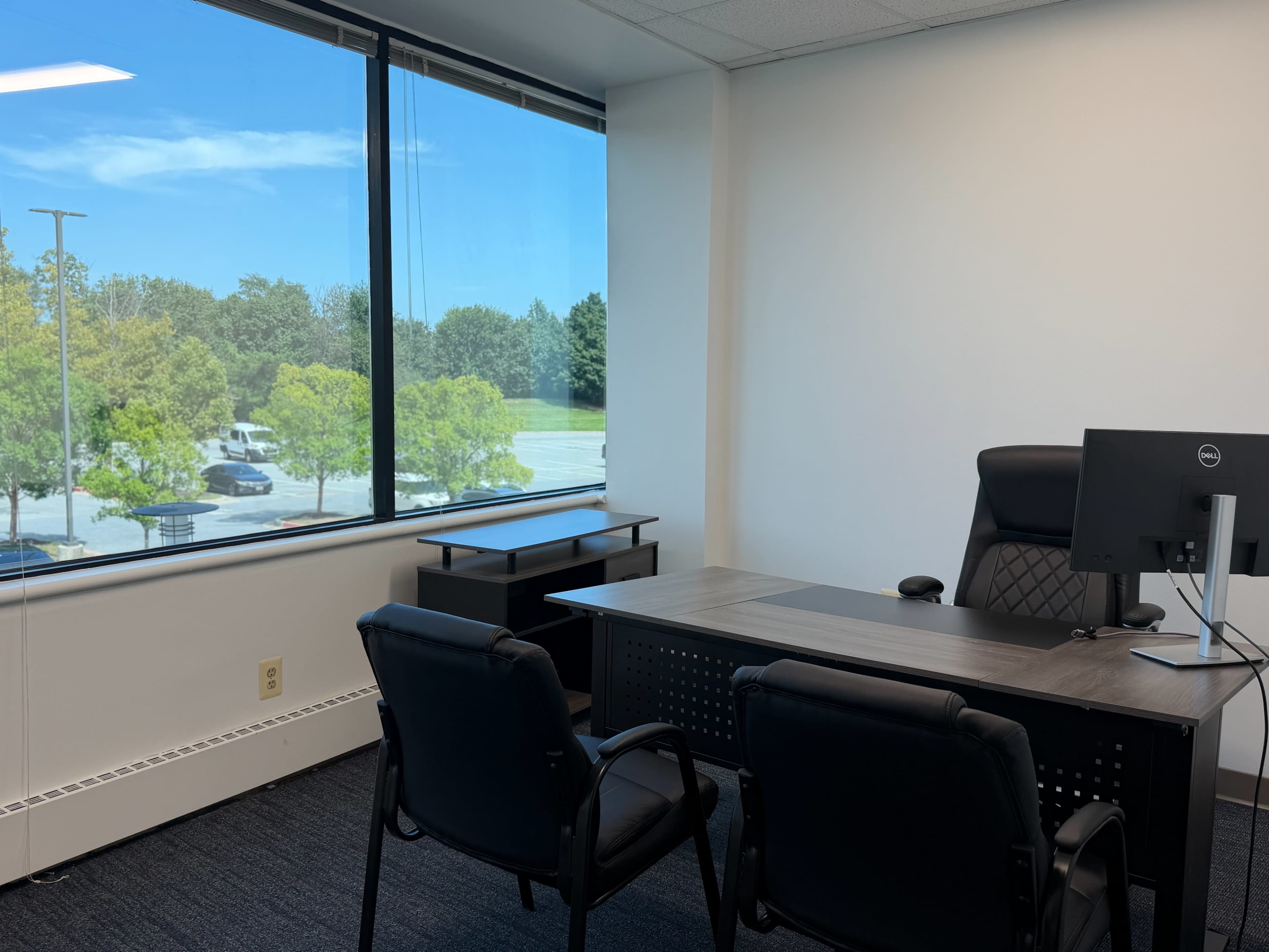 The image shows a modern office space with a desk, a computer monitor, two black chairs, and a large window revealing a view of trees and a parking lot outside.