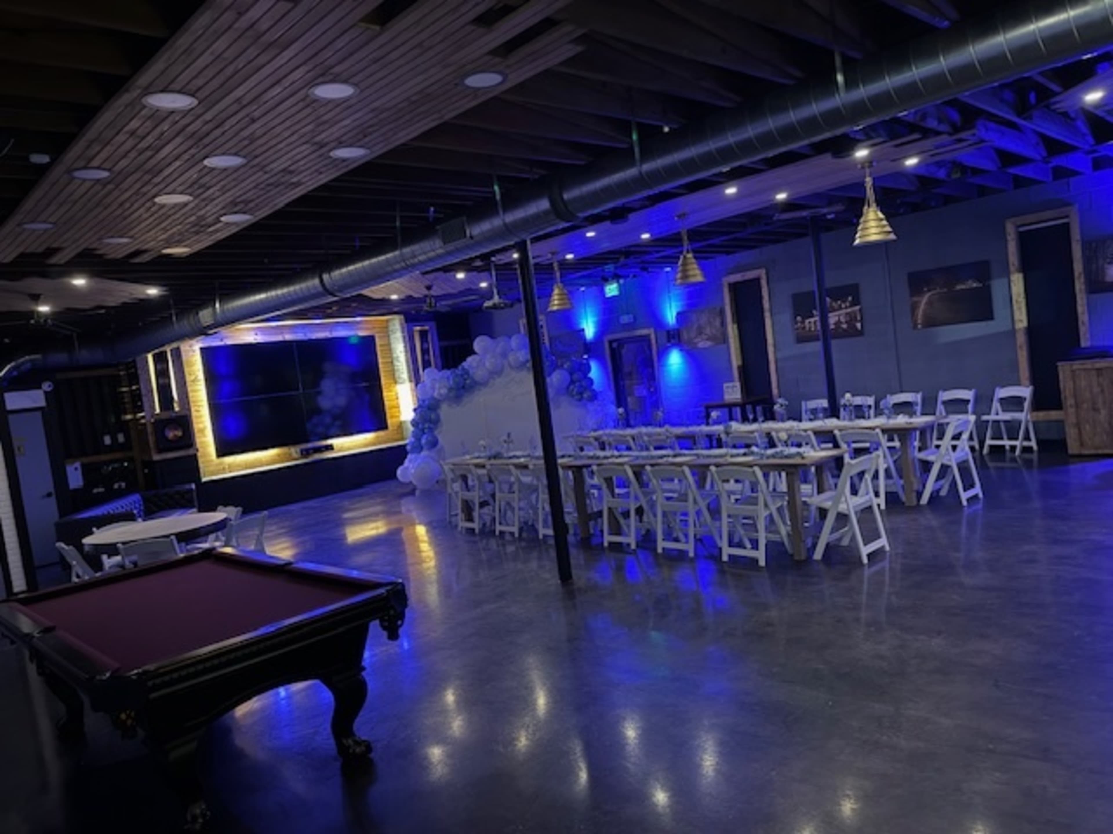 The image shows a spacious indoor venue with chairs and tables arranged for an event, featuring blue lighting, a large television screen, and decorative elements such as a balloon arch.
