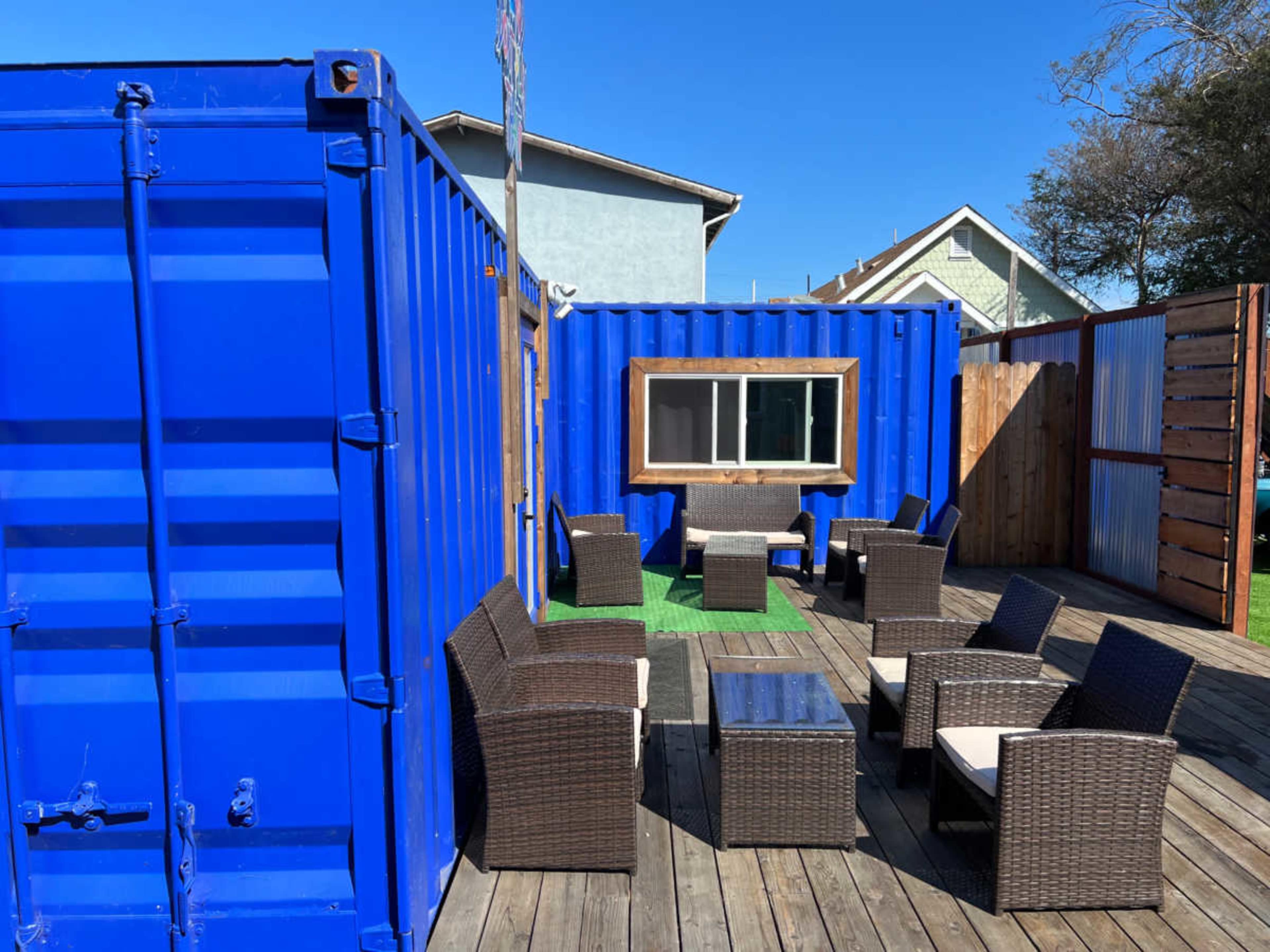 A seating area with wicker chairs and a table is arranged on a wooden deck, enclosed by blue shipping containers and wooden fencing.