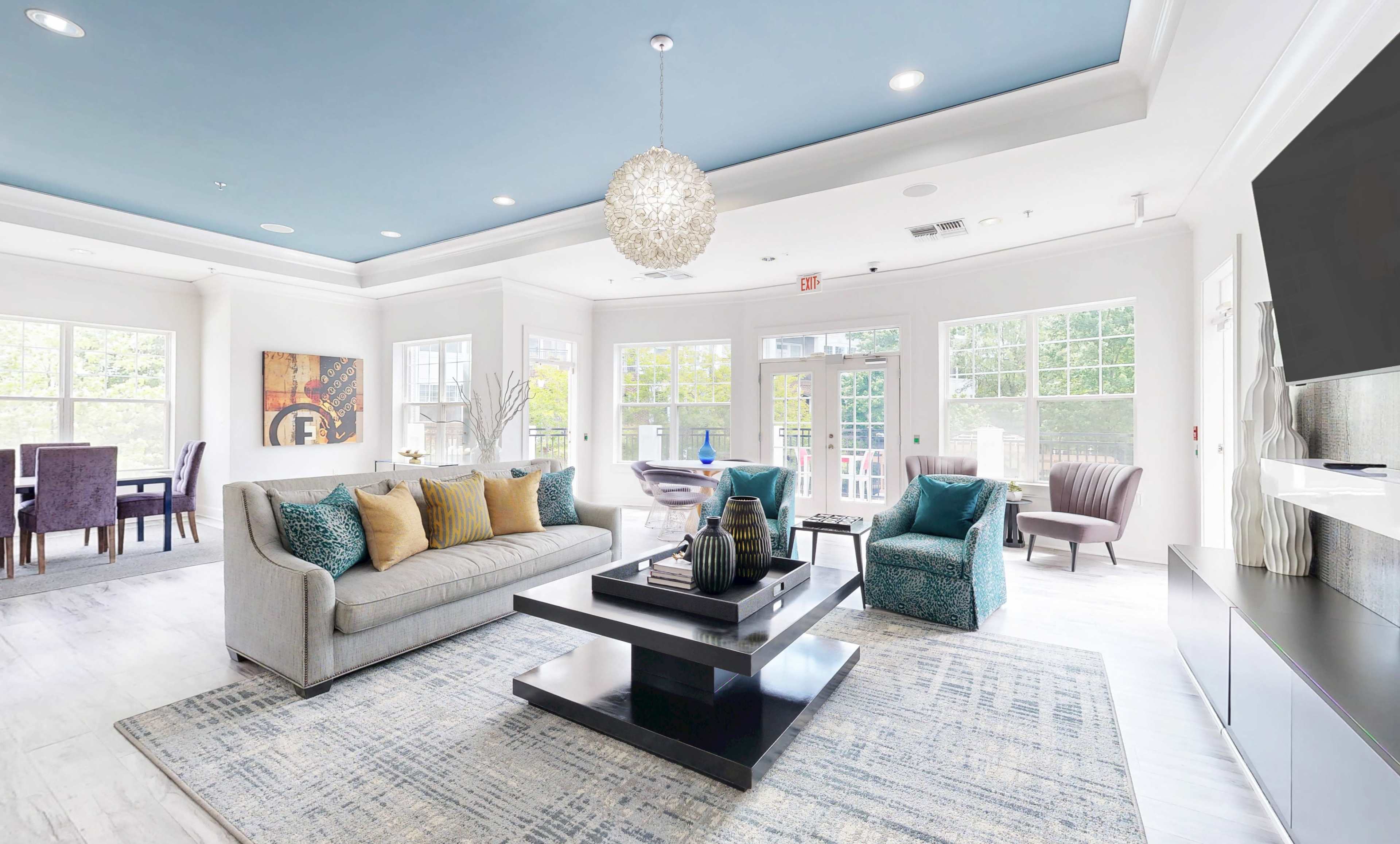 The image shows a modern living room with a gray sofa, decorative pillows, a coffee table, and a dining area with a table and chairs, all under a blue ceiling.
