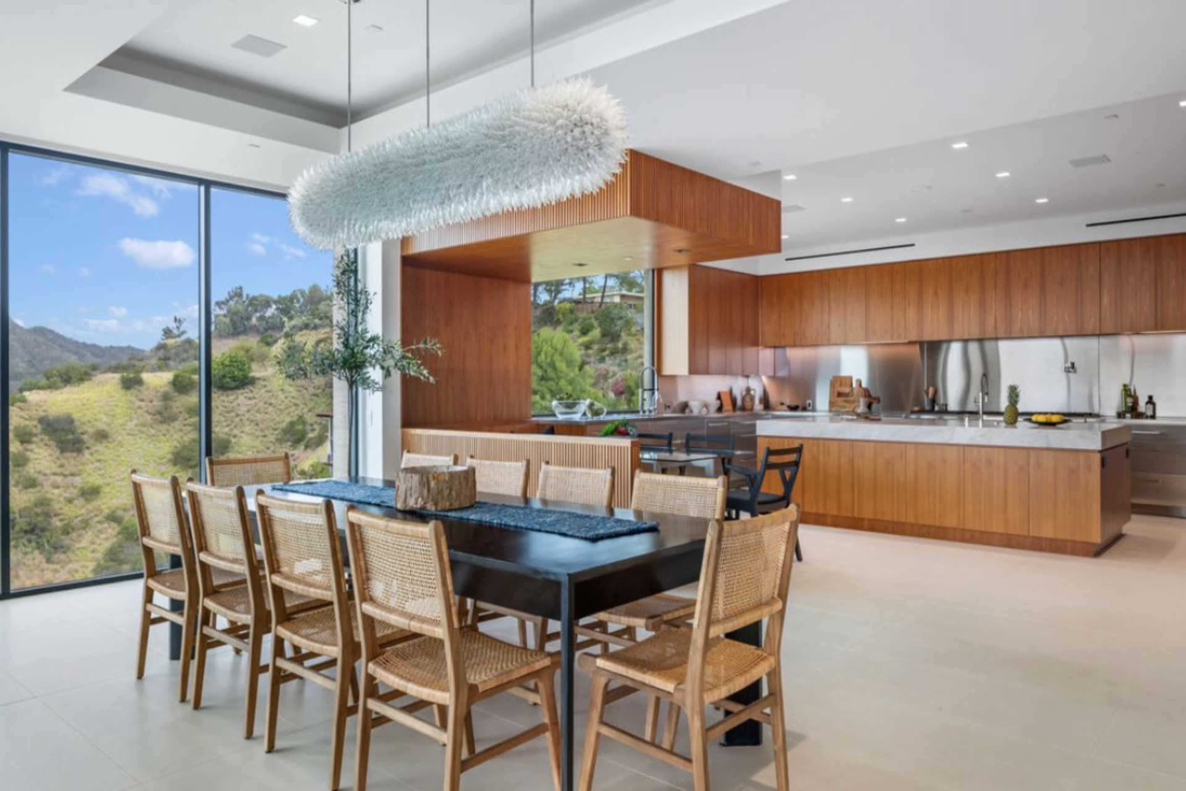A modern kitchen and dining area feature a wooden table with rattan chairs, a large window showcasing a hillside view, and sleek cabinetry.