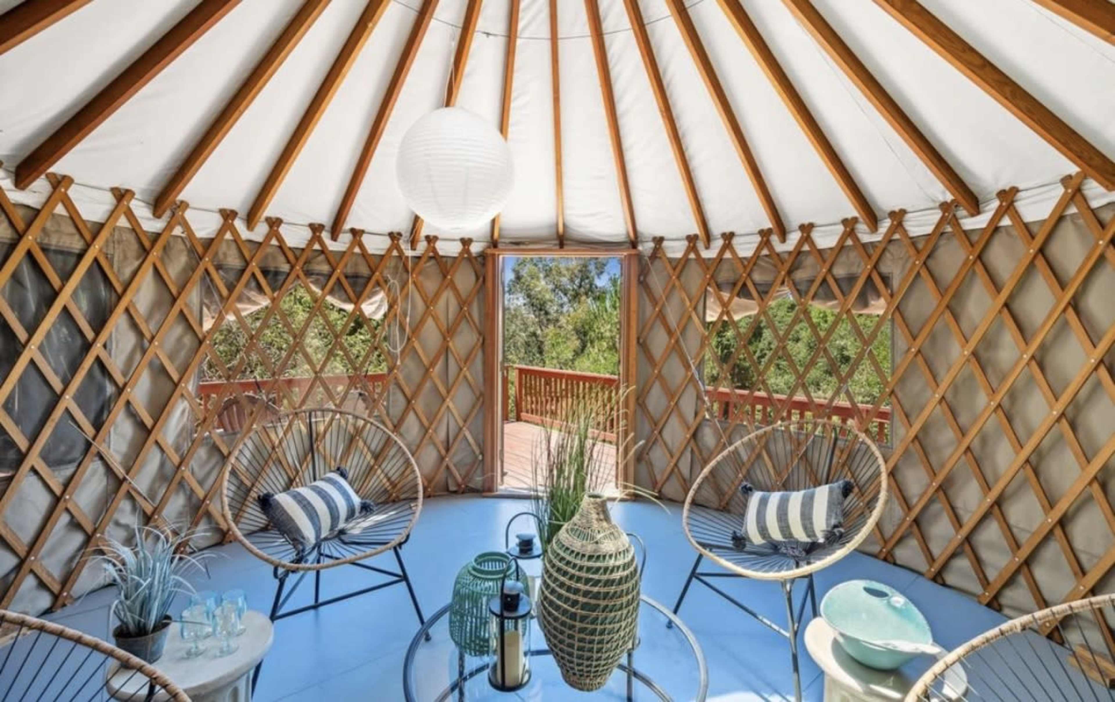 The interior of a yurt features wooden lattice walls, a round glass table, and two stylish chairs, all set against a backdrop of greenery visible through large windows.