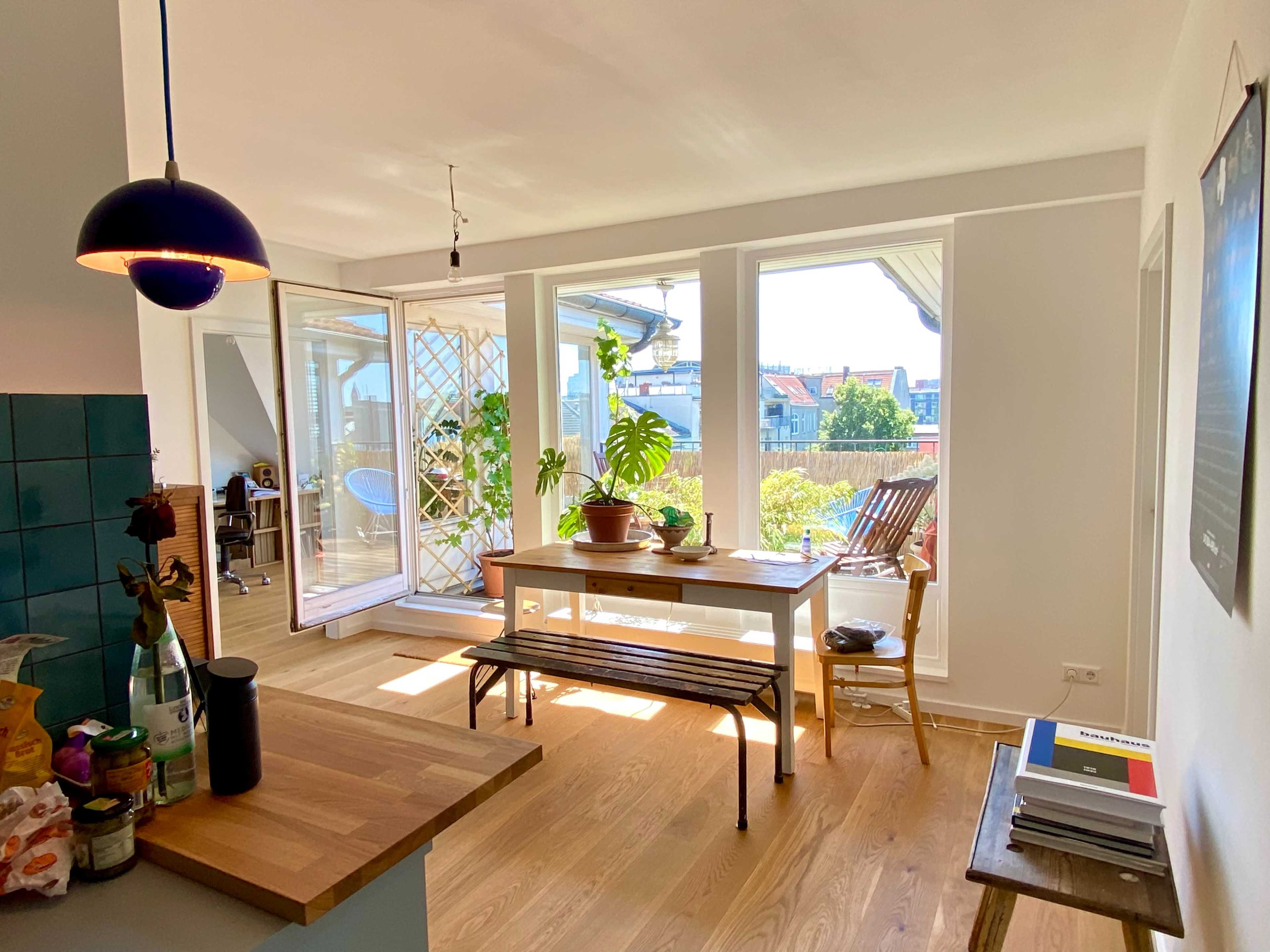 A bright, open kitchen and dining area features a wooden table surrounded by plants and large windows overlooking a balcony.