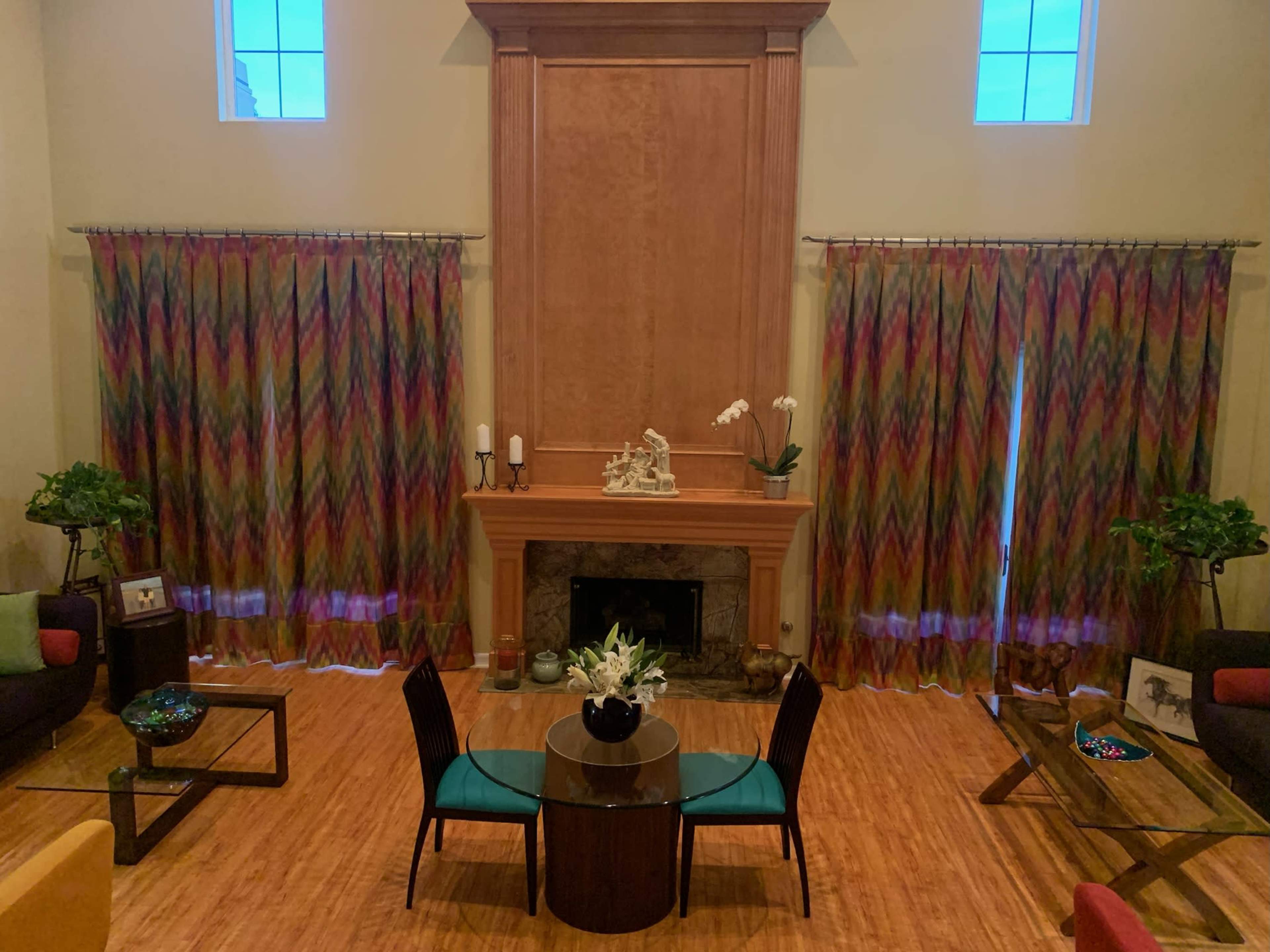 The image shows a spacious living room with a central round table, two chairs, a fireplace, and colorful curtains on either side of two windows.
