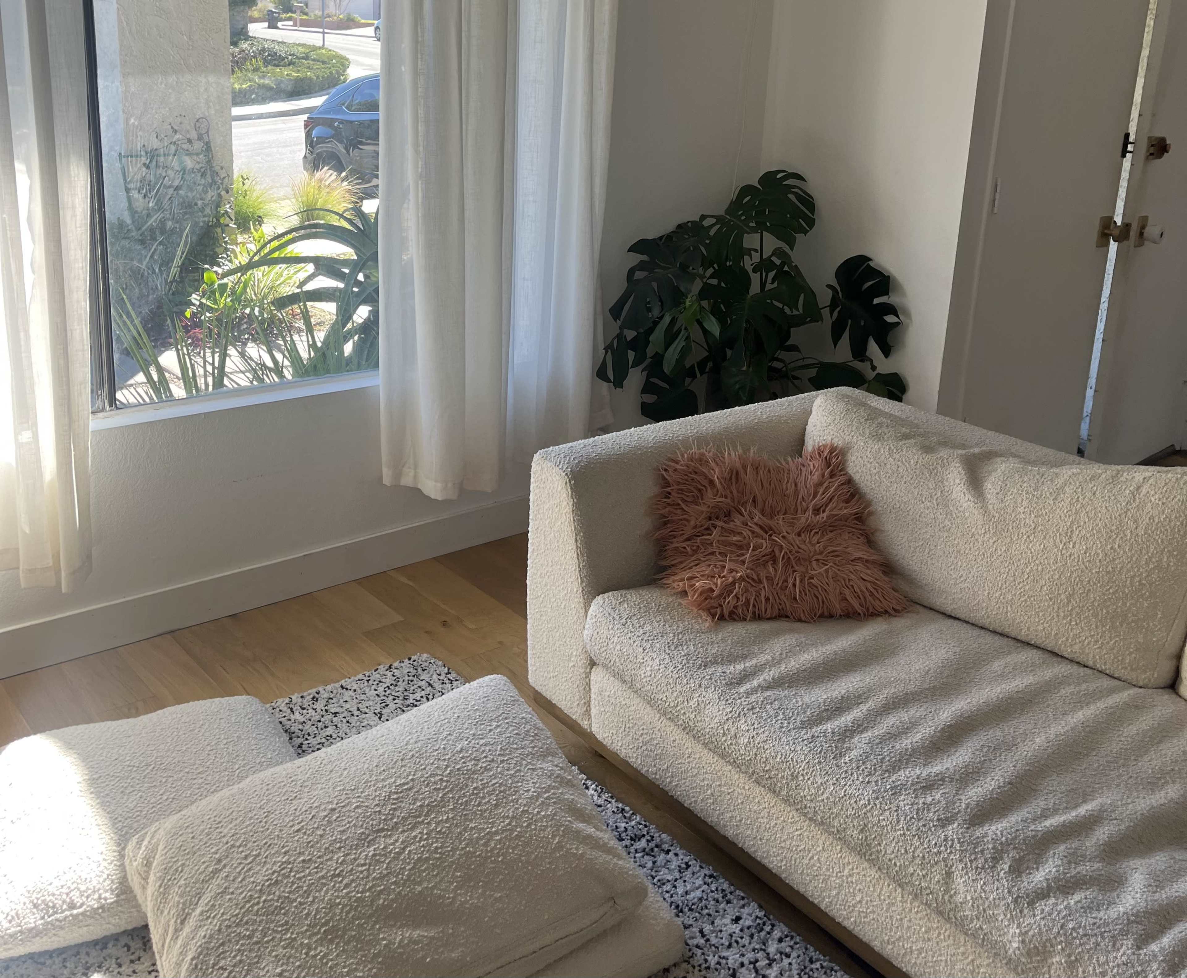 A cozy living room with a white sofa, a plush pink pillow, and sunlight streaming through sheer curtains onto a wooden floor.