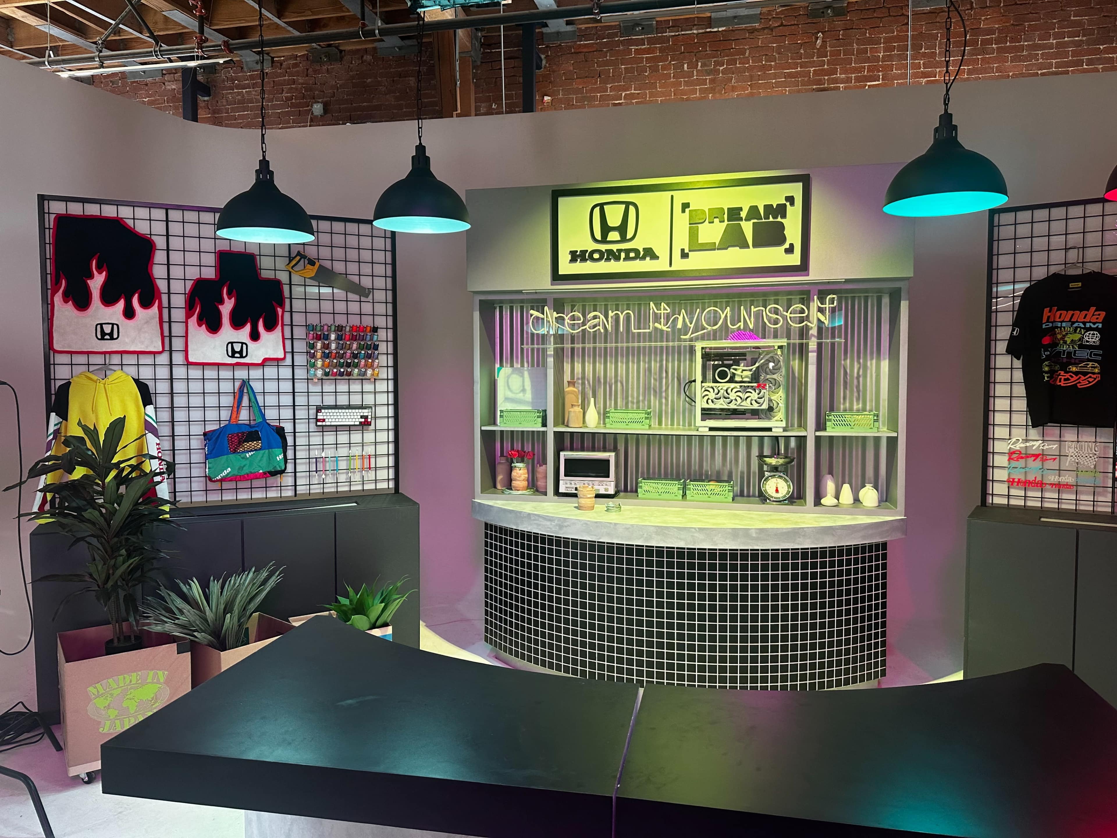 The image shows a colorful promotional space featuring Honda branding, including merchandise displays, a counter, and neon signage that reads "dream it, try yourself."