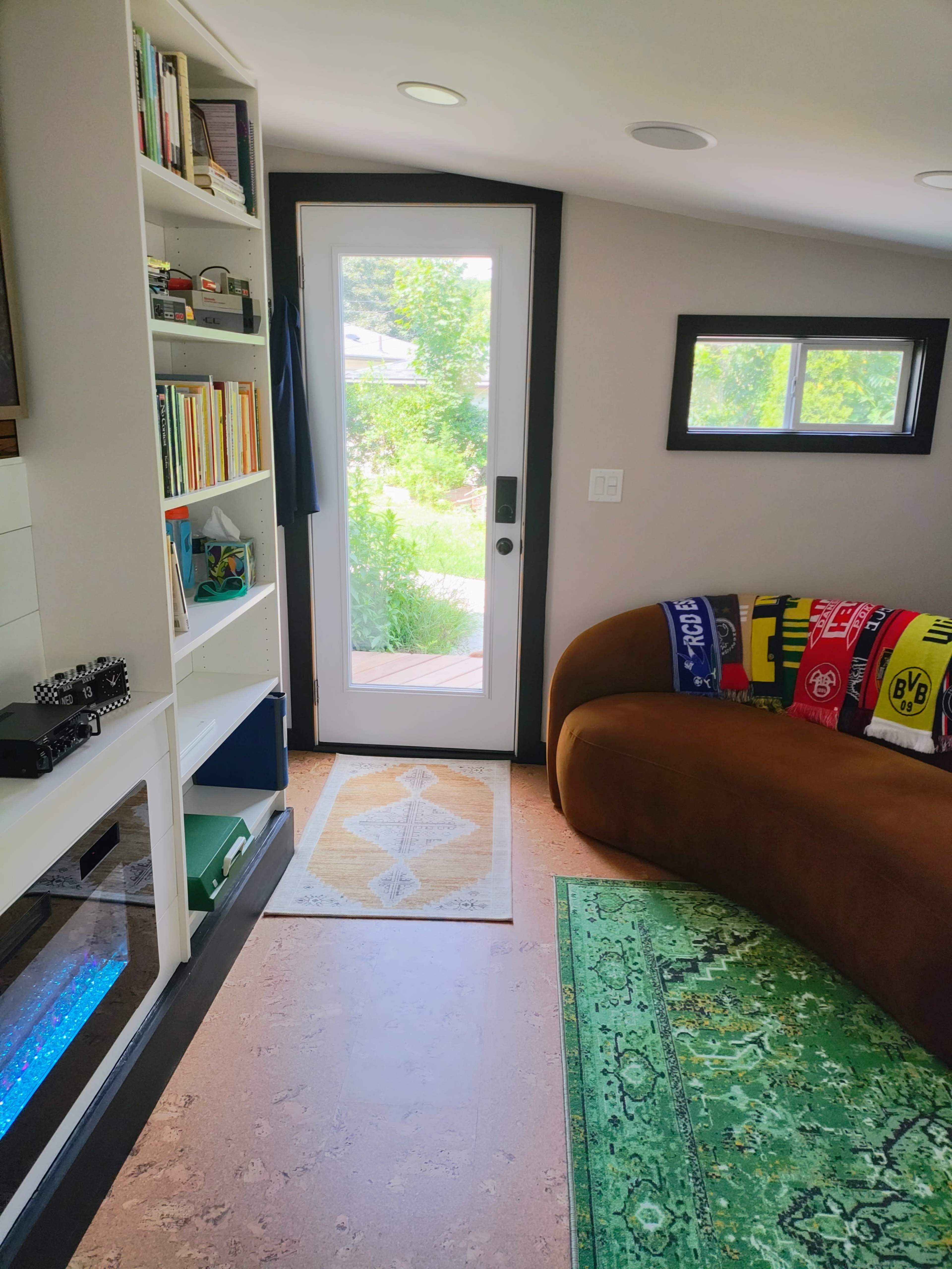 The image shows a cozy interior space featuring a curved brown couch, bookshelves, and a doorway leading to an outdoor view.
