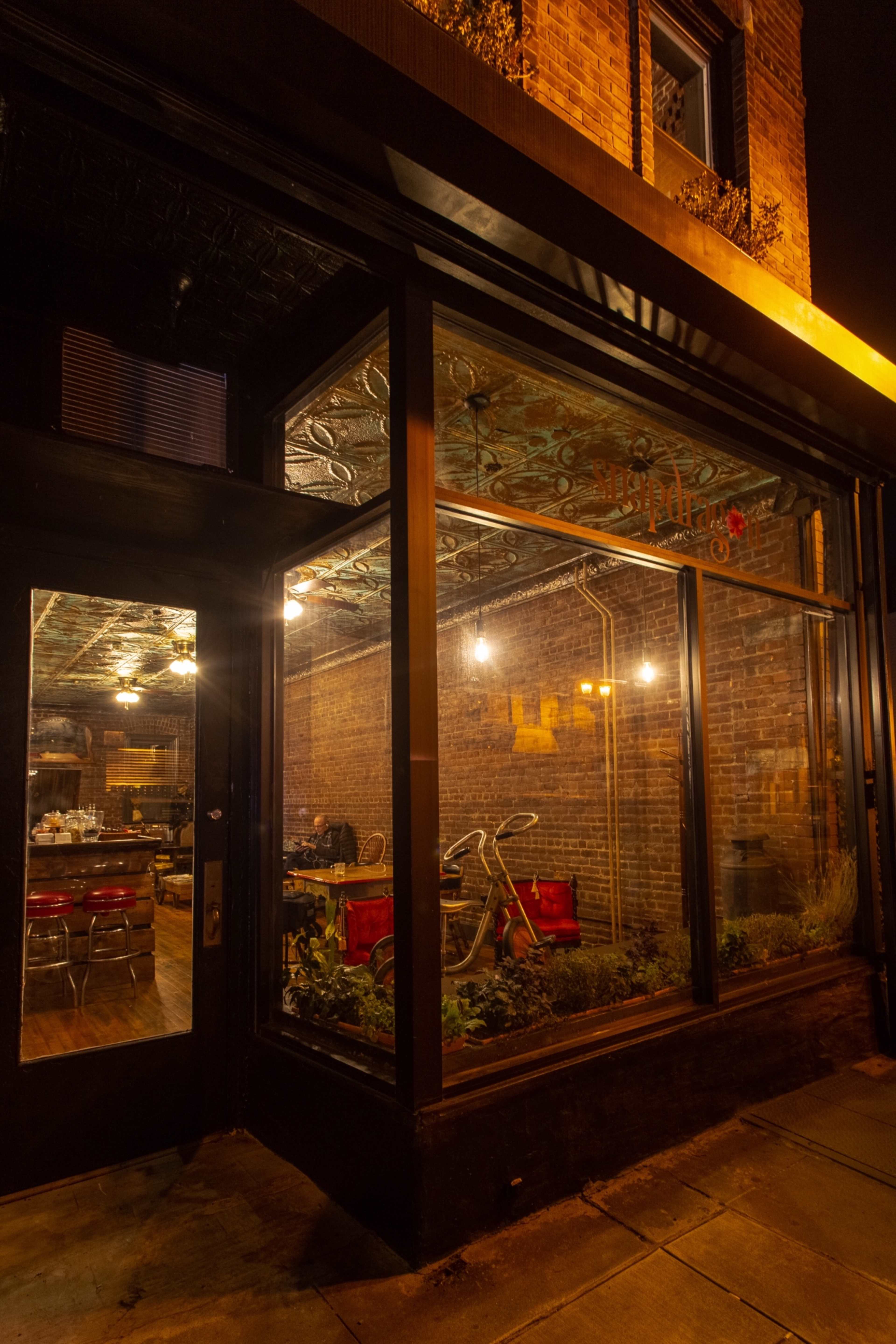 The exterior of a restaurant with large windows, revealing an interior with brick walls and red seating, illuminated by pendant lights at night.