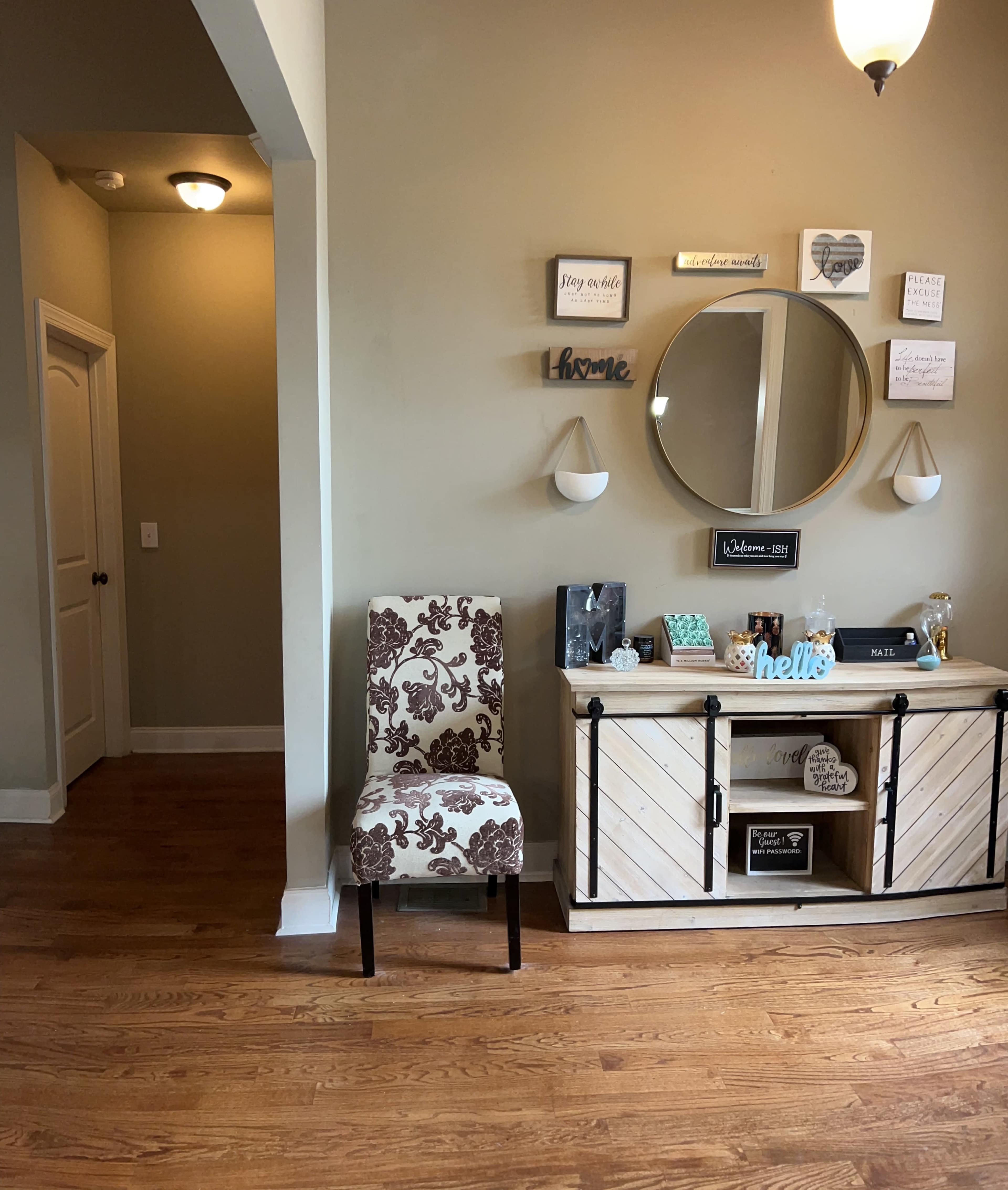 The image shows a well-decorated entryway with a wooden console table, a circular mirror, decorative wall signs, and a patterned chair.