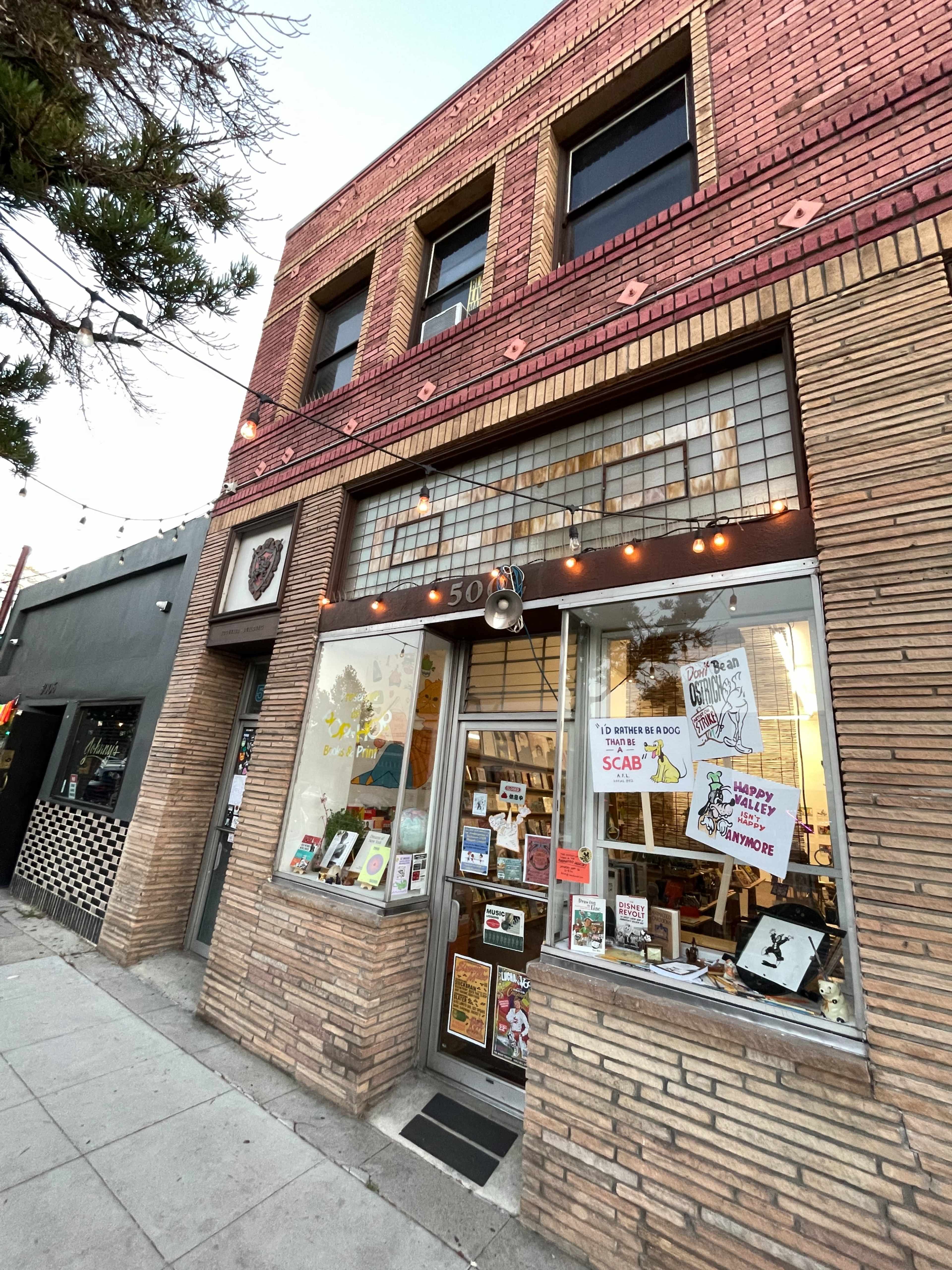 The image shows a storefront with large windows displaying various art and craft items, set within a brick building featuring a red and beige facade.