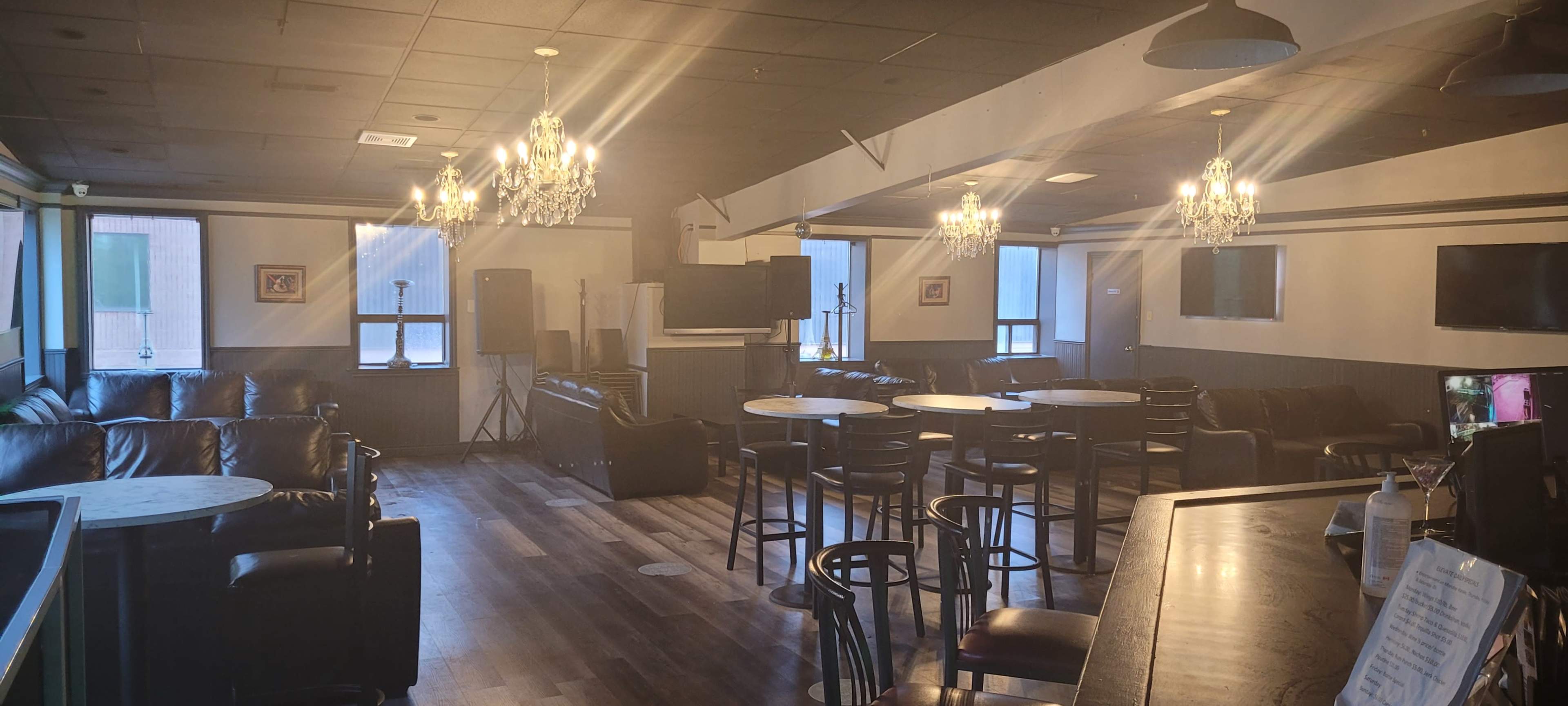 A spacious lounge area features leather seating, circular tables, and decorative chandeliers, with large windows allowing natural light to enter.