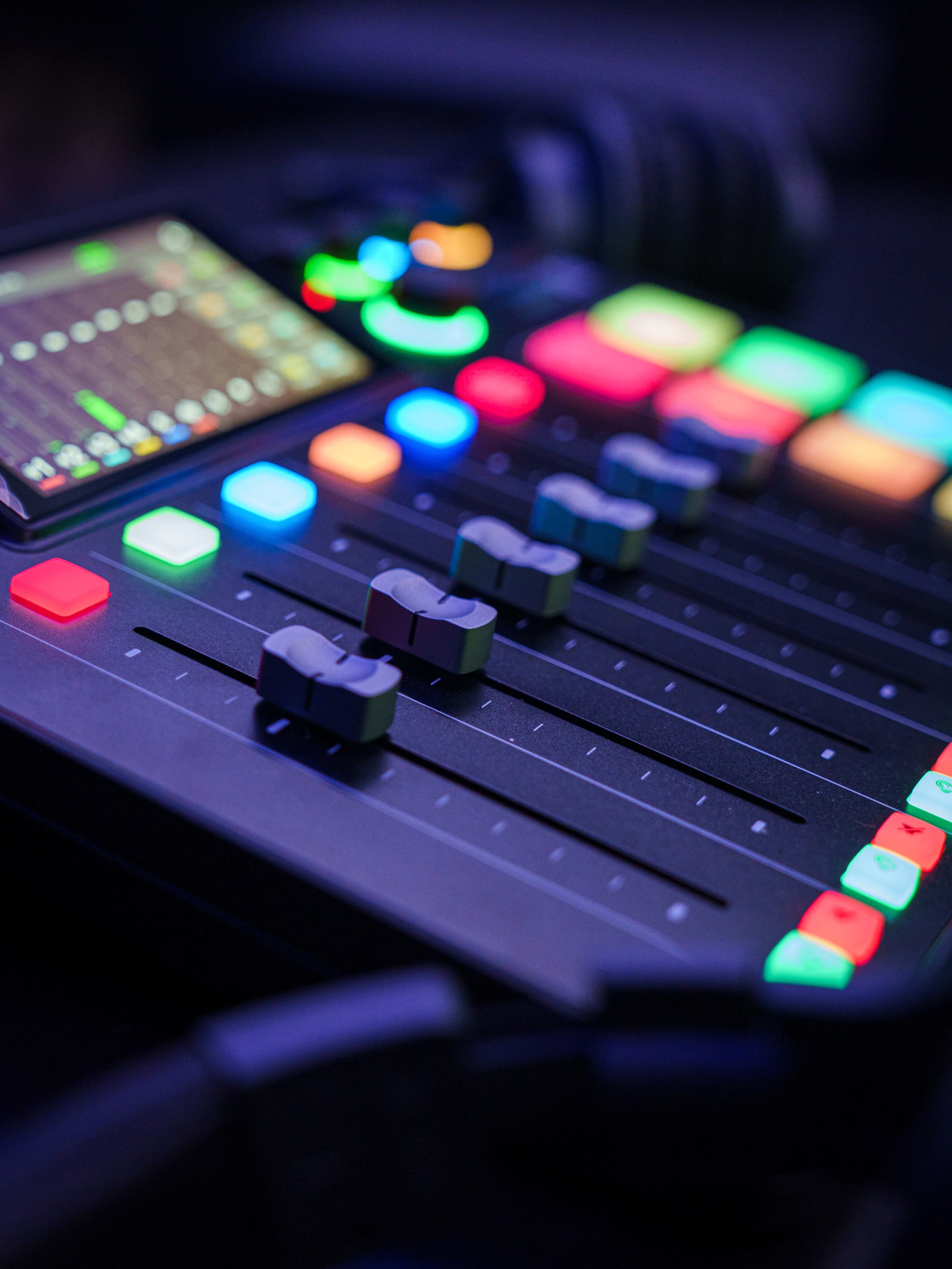 A digital audio mixing console with illuminated faders and buttons is displayed on a dark surface.