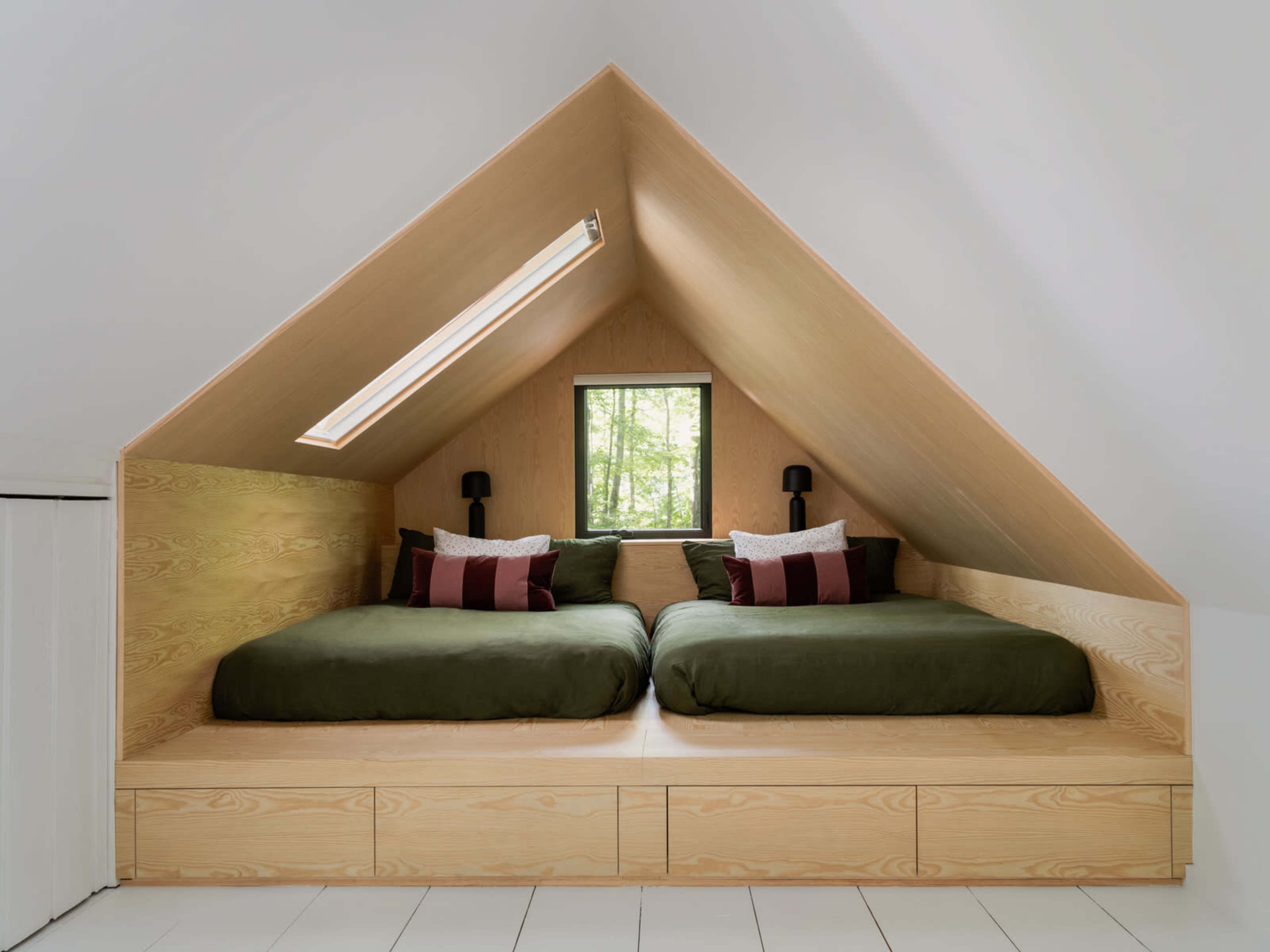 The image shows an attic bedroom with two green beds placed on a wooden platform under a sloped ceiling, featuring a window that overlooks trees.