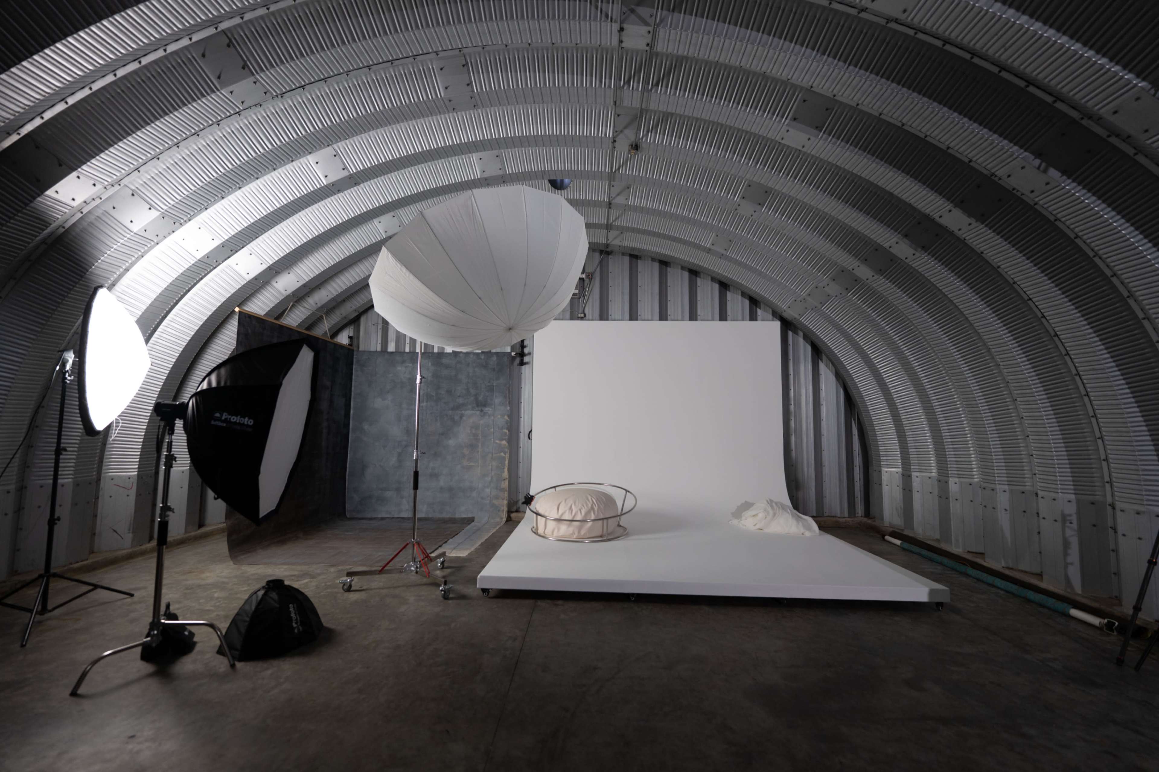 The image shows a photography studio with a curved metal ceiling, featuring a backdrop, lighting equipment, and a minimalist setup with props on a platform.