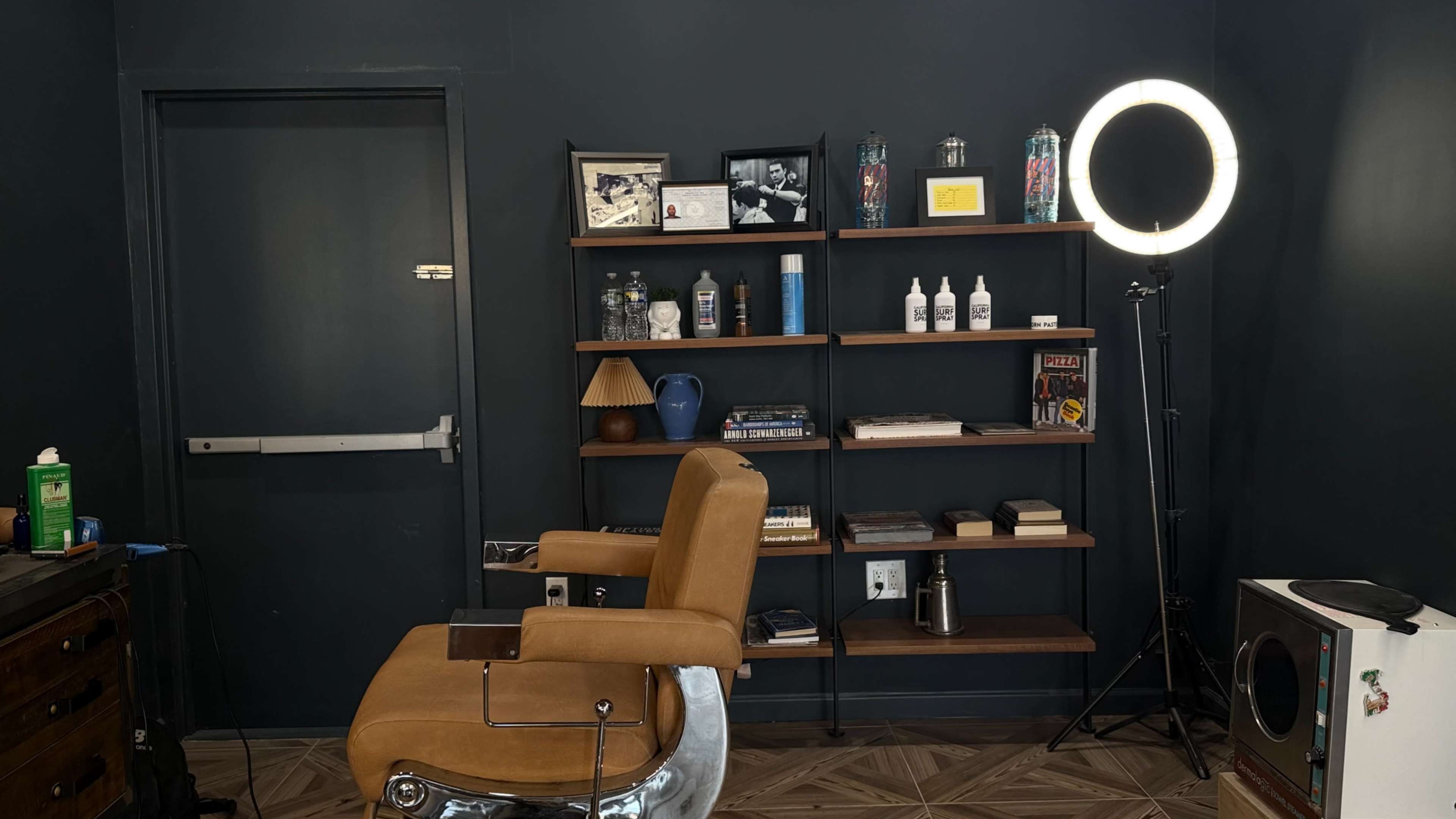 The image shows a barber chair in front of a dark wall, with a shelf holding various decorative items and products, and a ring light positioned nearby.