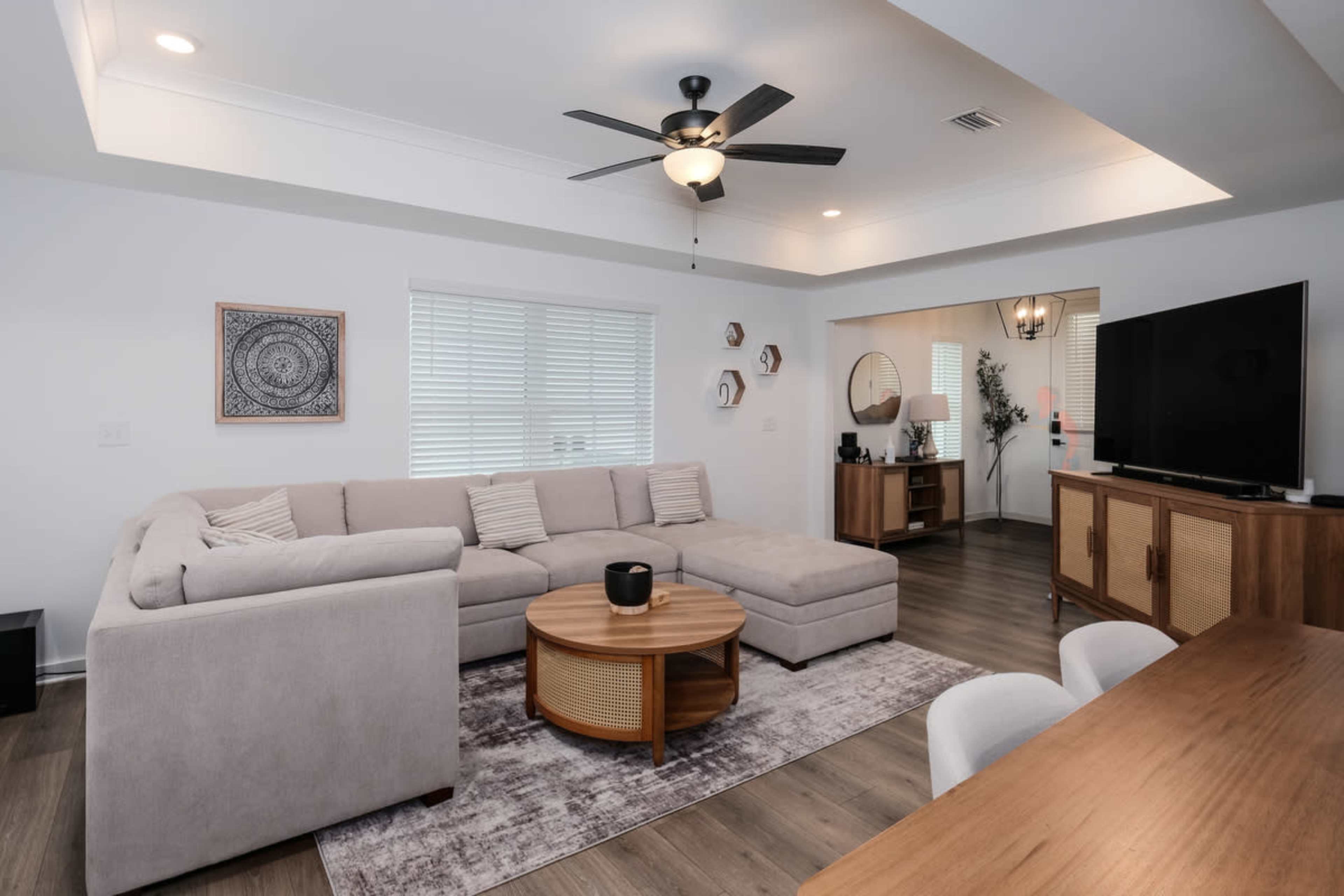 The image shows a modern living room featuring a large sectional couch, a round coffee table, and a television on a wooden stand, with light-colored walls and hardwood flooring.