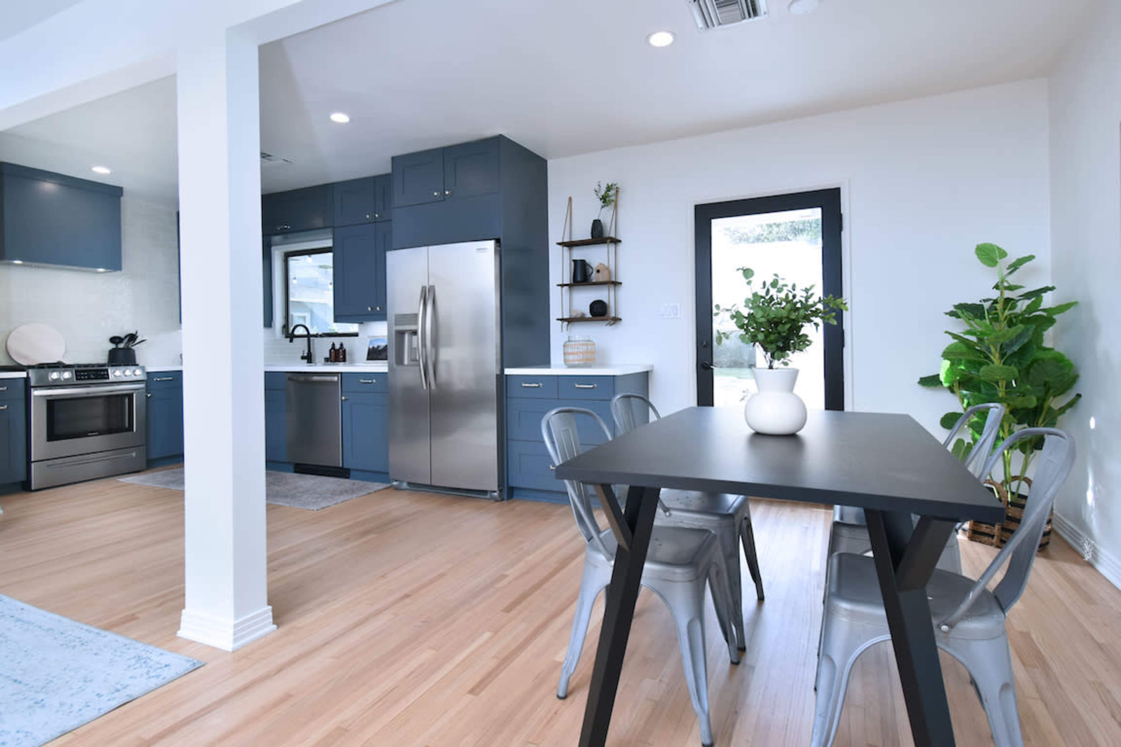 The image depicts a modern kitchen and dining area with blue cabinetry, stainless steel appliances, a wooden dining table, and a potted plant.
