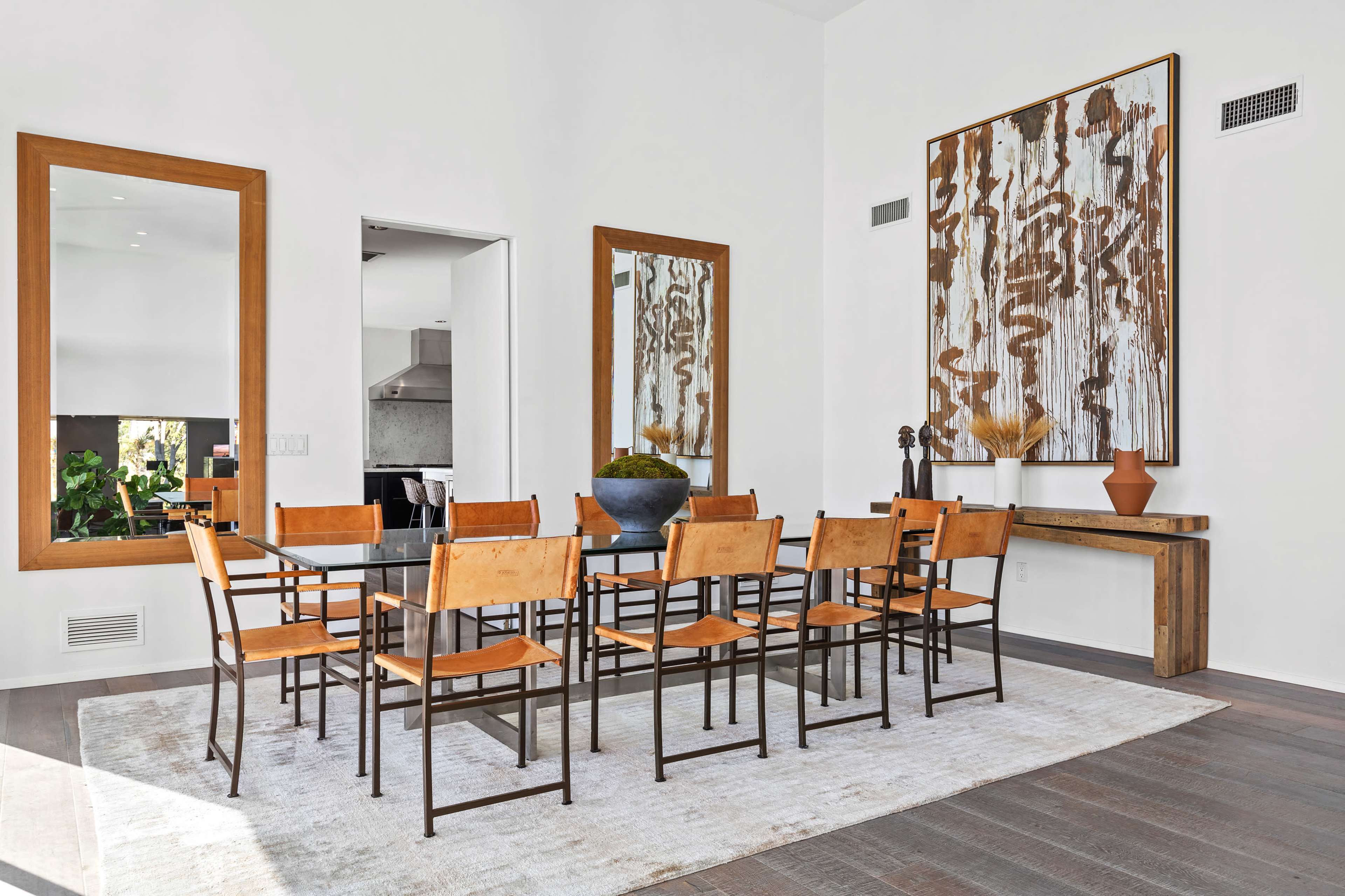A dining room features a large table surrounded by metal and wood chairs, with two mirrors and a piece of artwork on the walls.