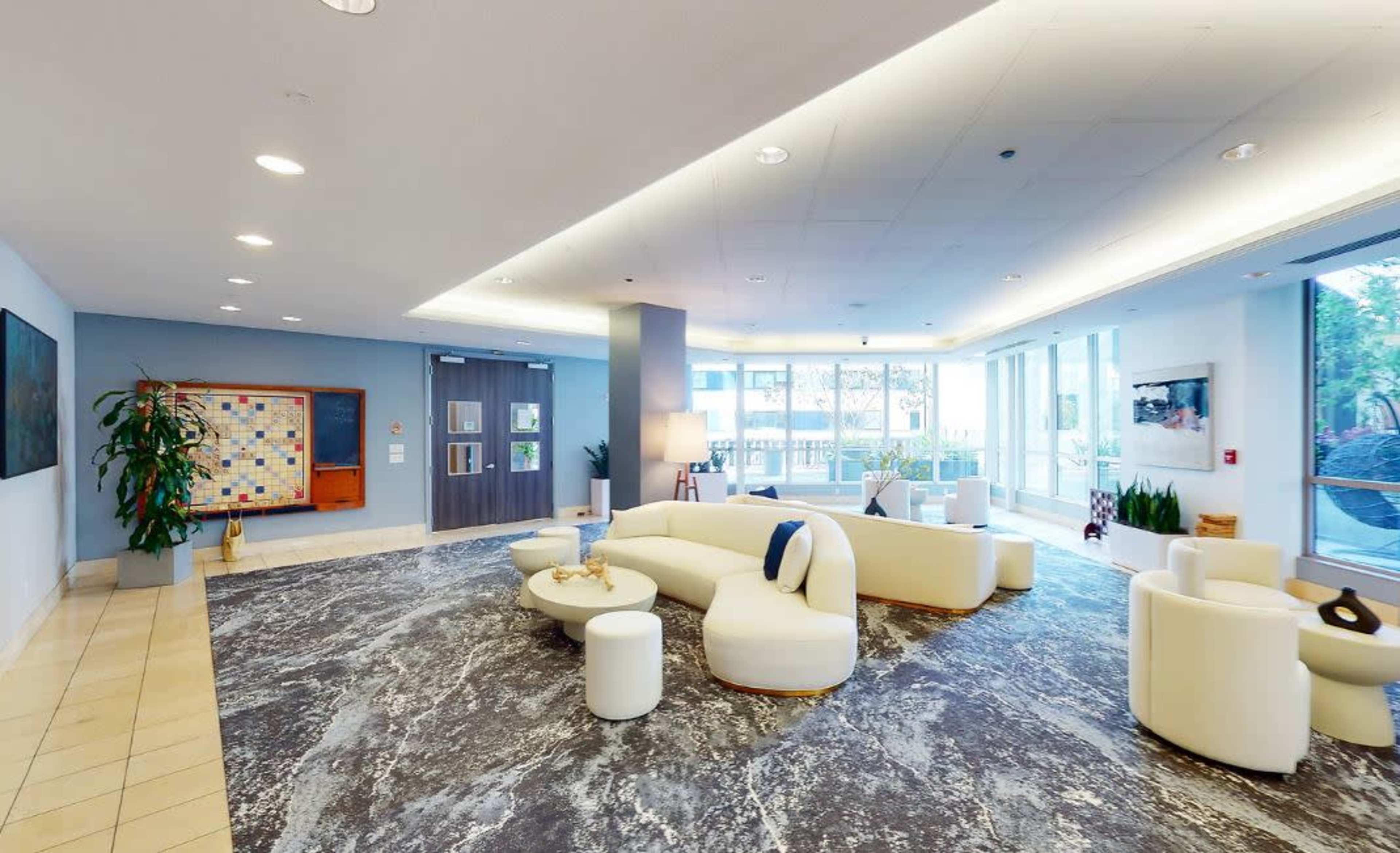 The image shows a modern lobby area featuring a large white curved sofa, small tables, a textured gray carpet, and floor-to-ceiling windows providing ample natural light.