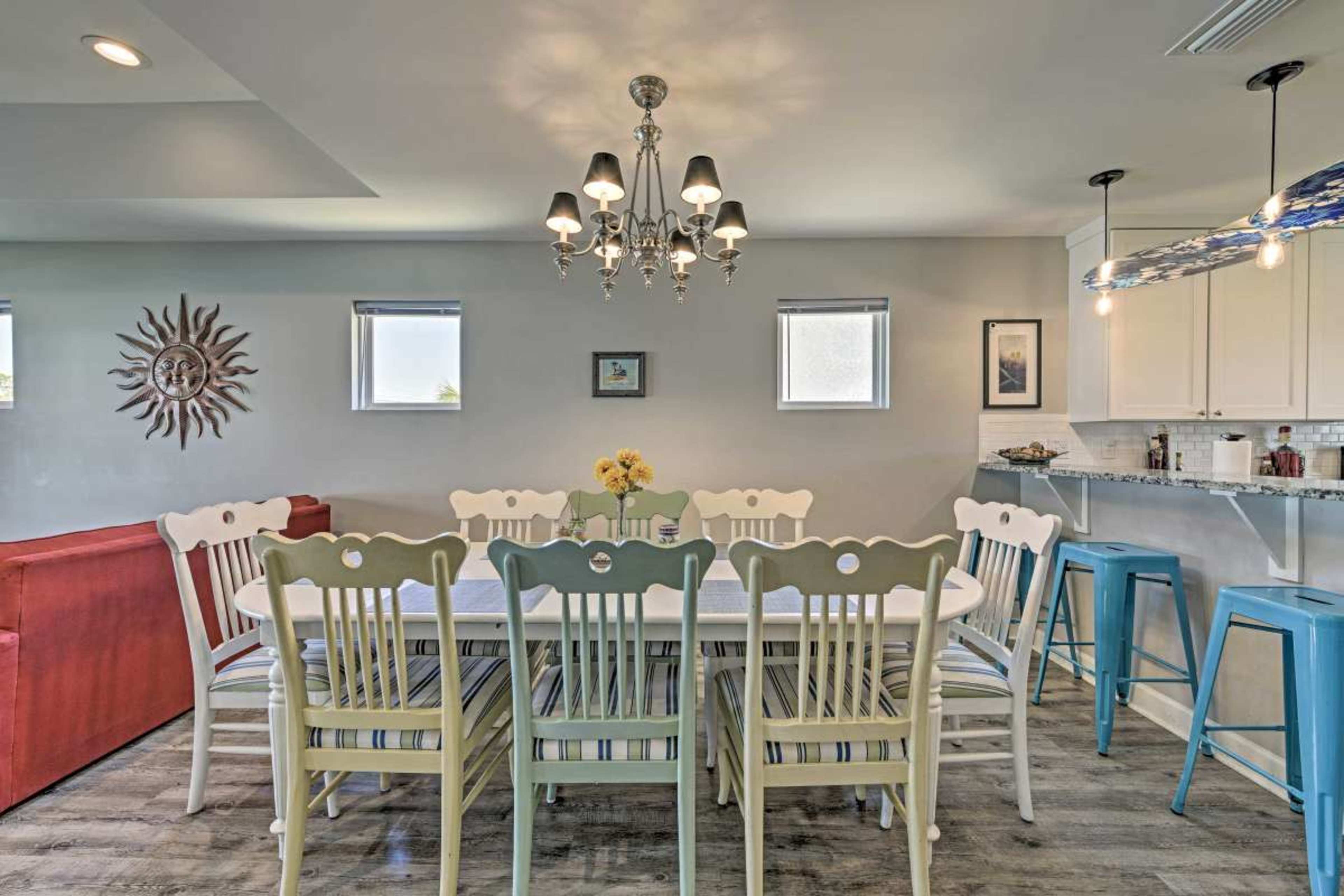 A large dining table surrounded by white wooden chairs is set in a well-lit room with a chandelier and modern bar stools in the background.