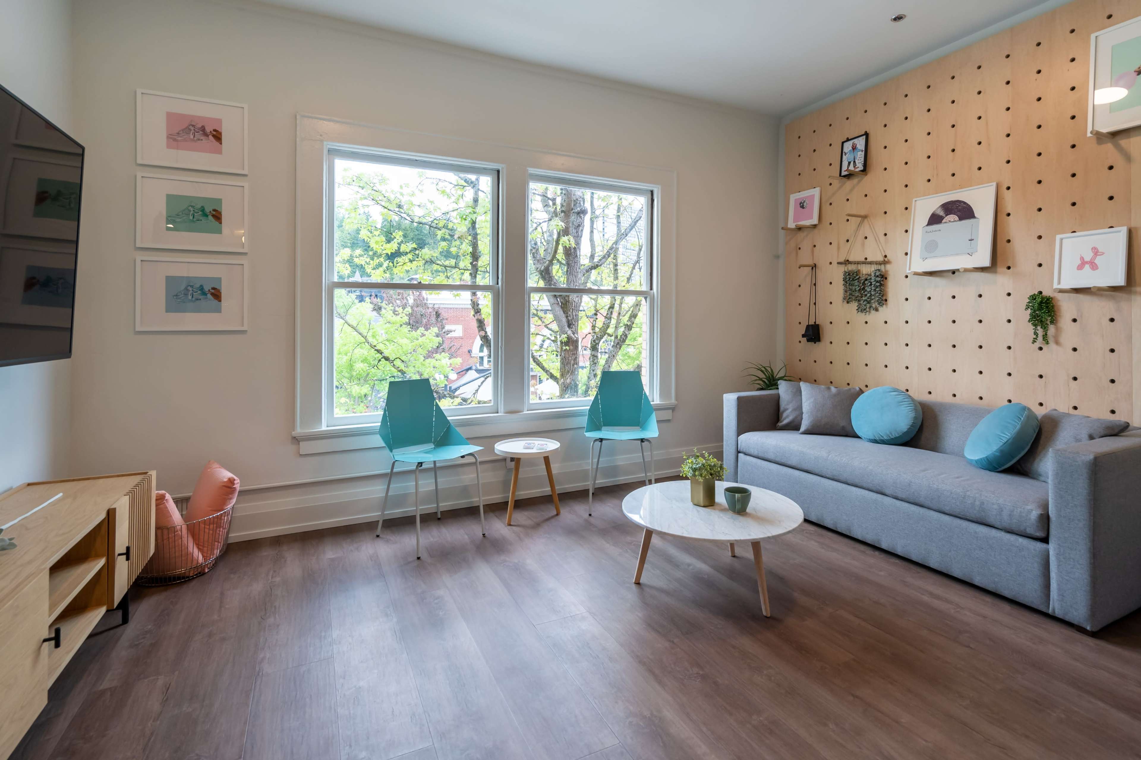 A modern living room features a gray sofa, teal chairs, a round coffee table, and large windows that frame a view of greenery outside.