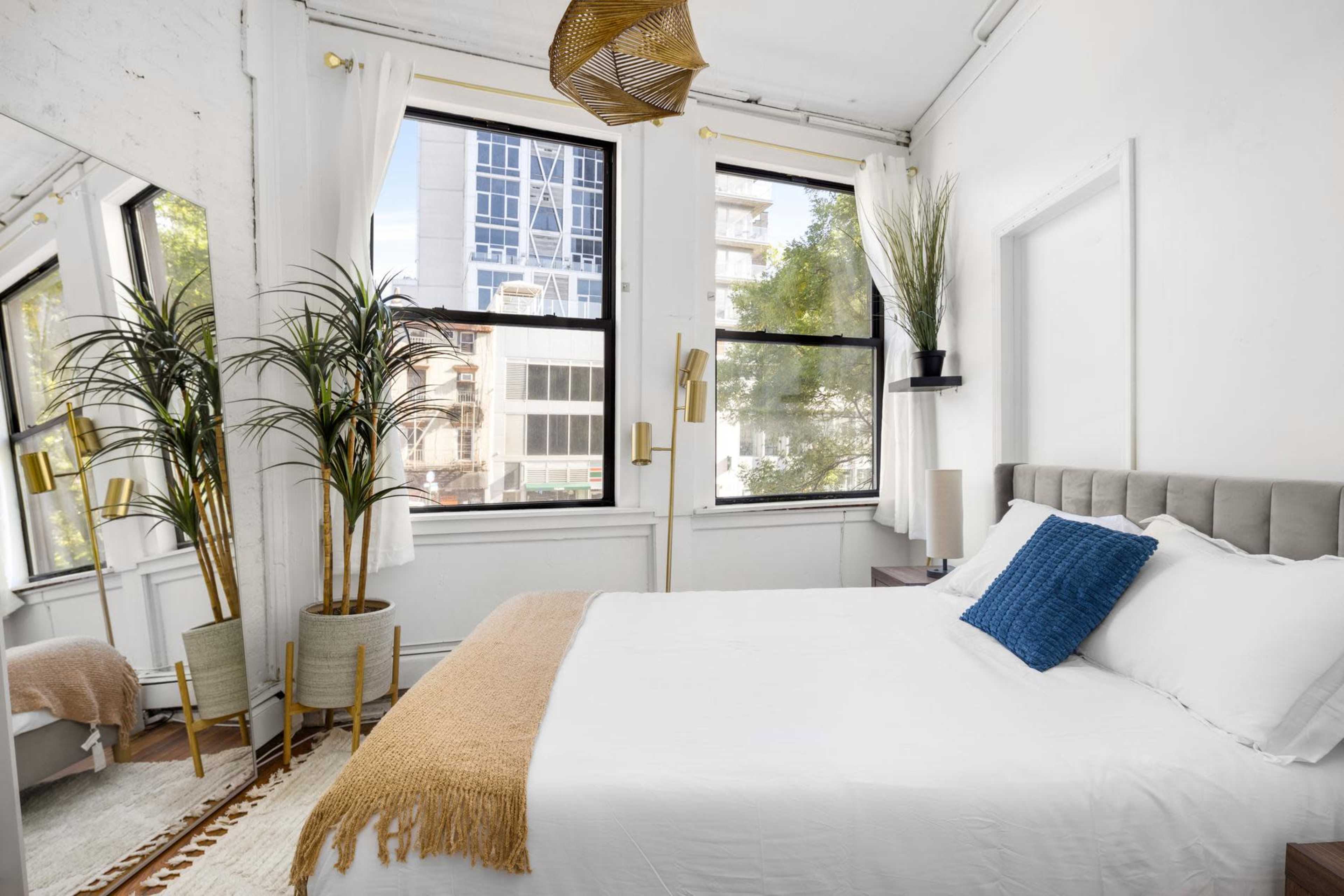 A bright bedroom features a bed with white linens, a blue throw pillow, large windows with city views, and decorative plants.