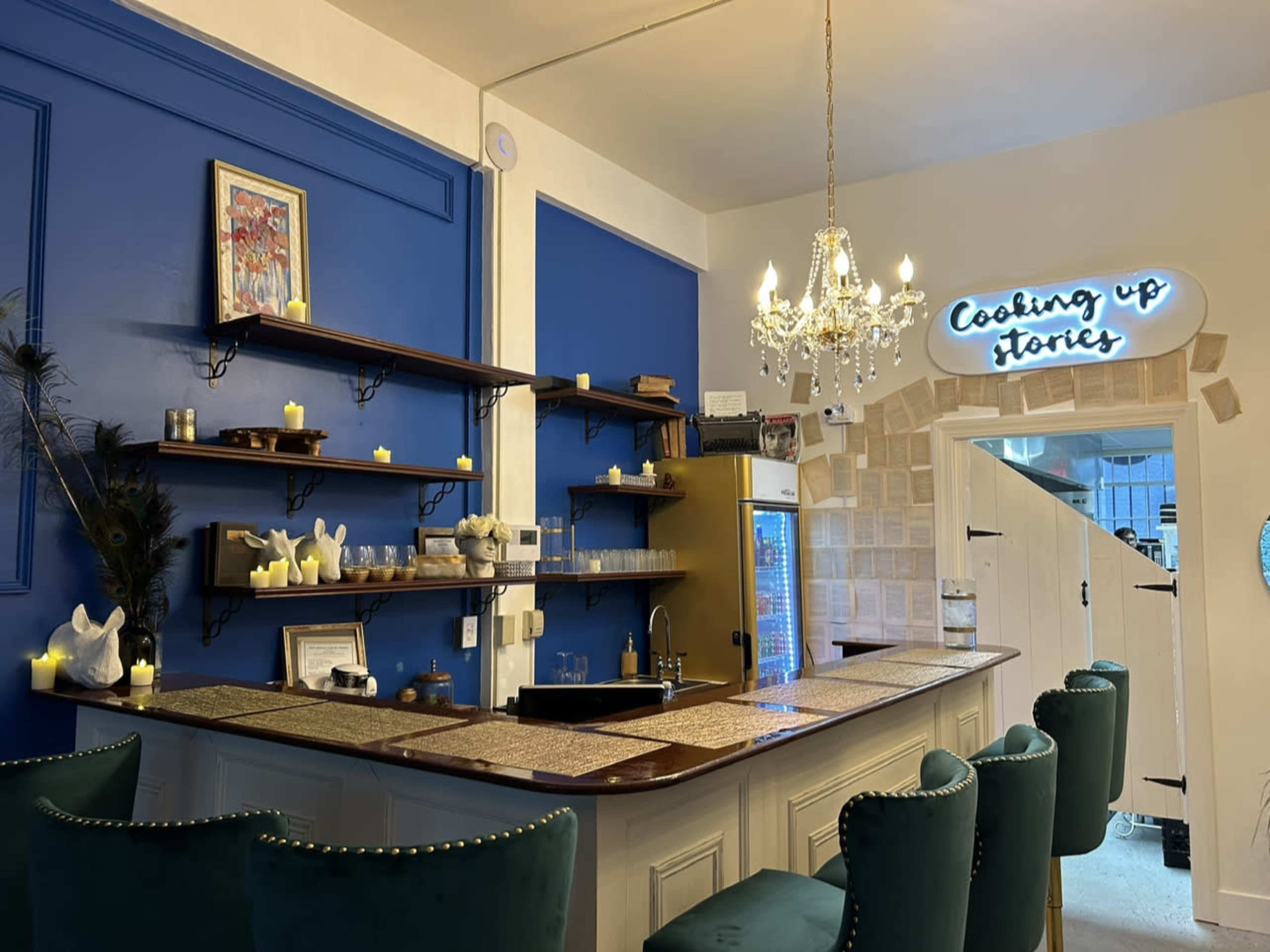 A stylish bar area with a blue wall, shelves displaying decorative items, a chandelier, and a illuminated sign that reads "Cooking up stories."