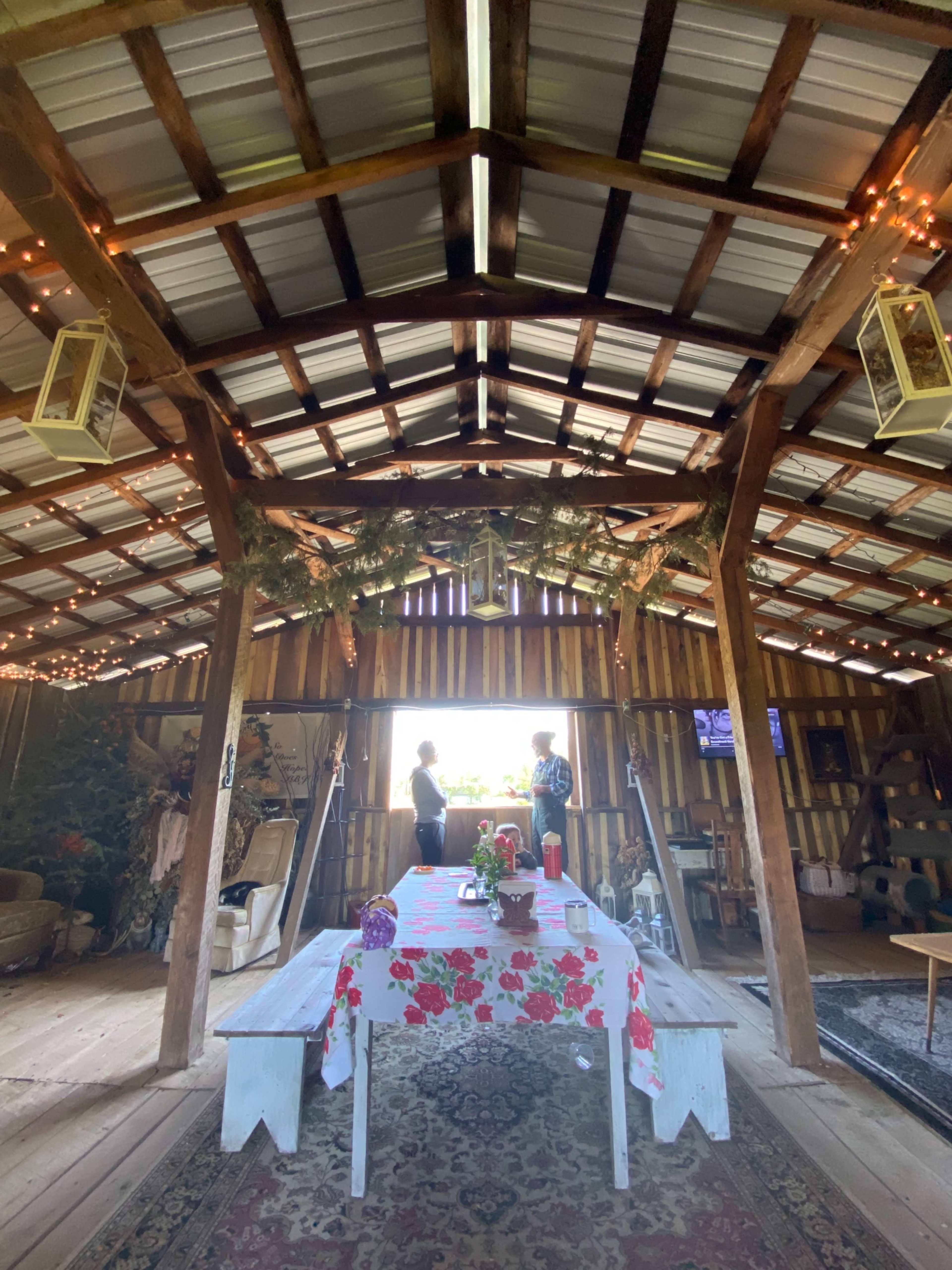 The interior of a rustic barn features a table with a floral tablecloth and two people standing by a window, surrounded by wooden beams and decorative lights.