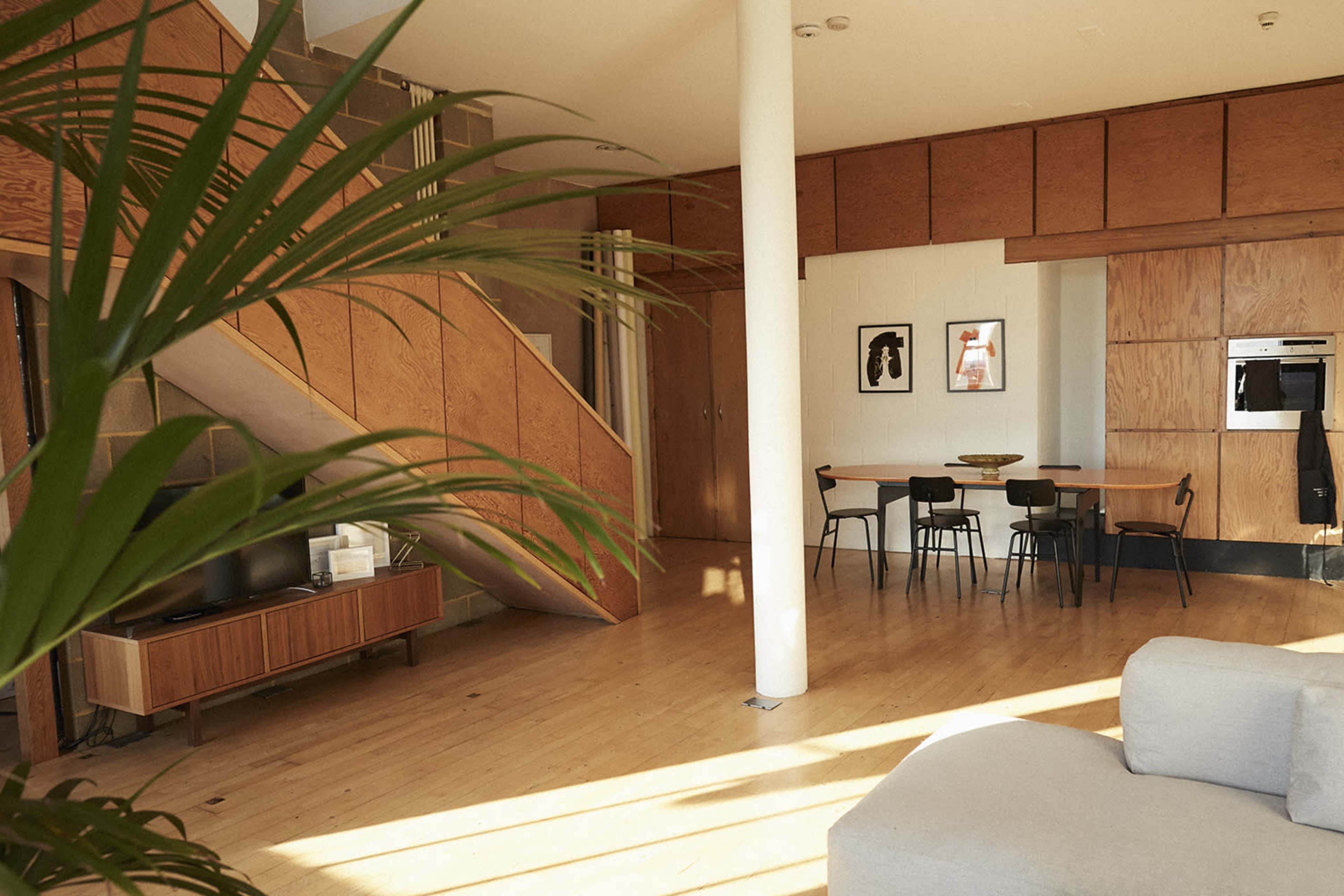 A modern living space with a minimalist design featuring a staircase, wooden walls, a dining area with a round table, and a seating area with a sofa.